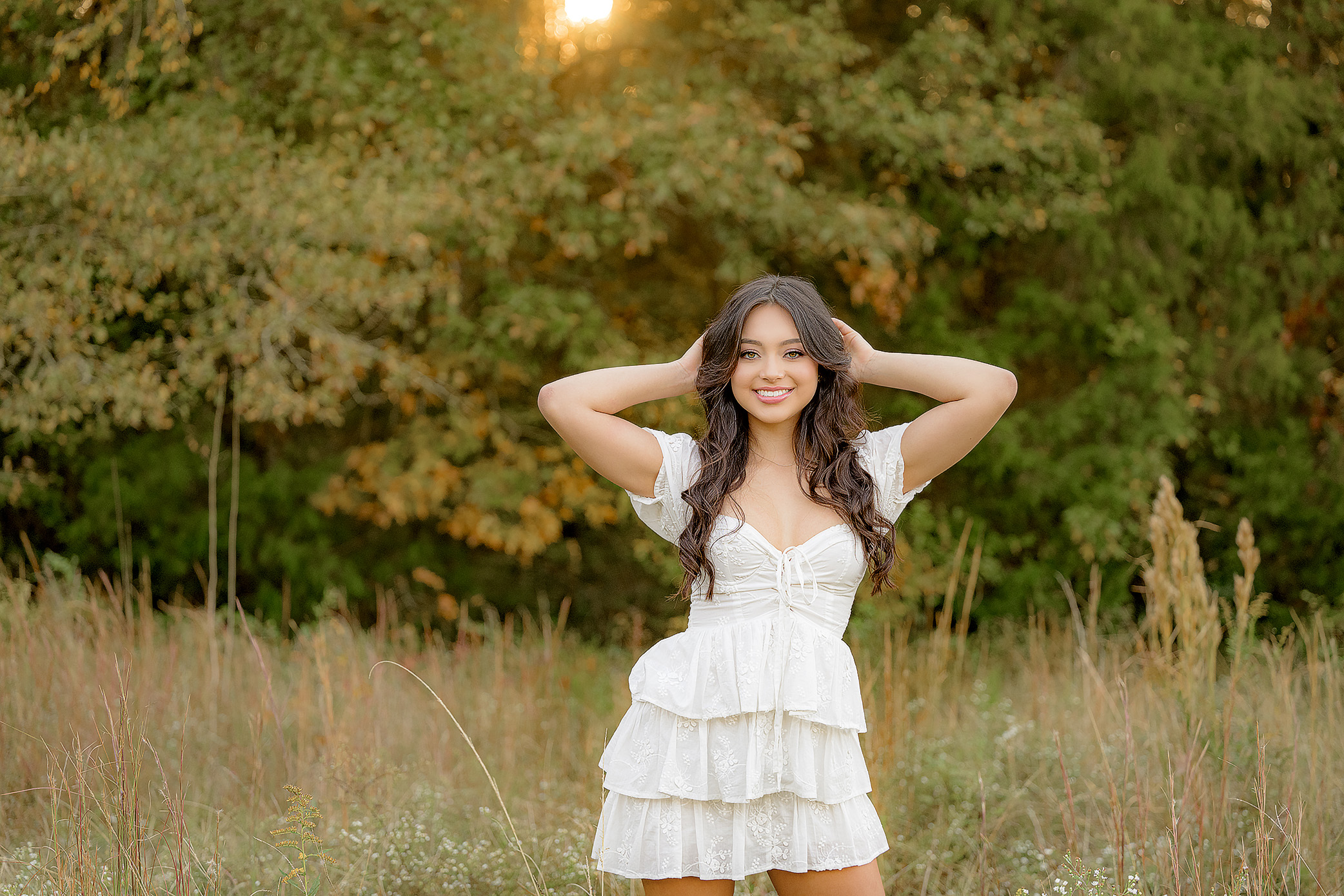 Senior and class of 2025 graduate smiles at the camera with her hands playfully in her hair, dressed in a white dress