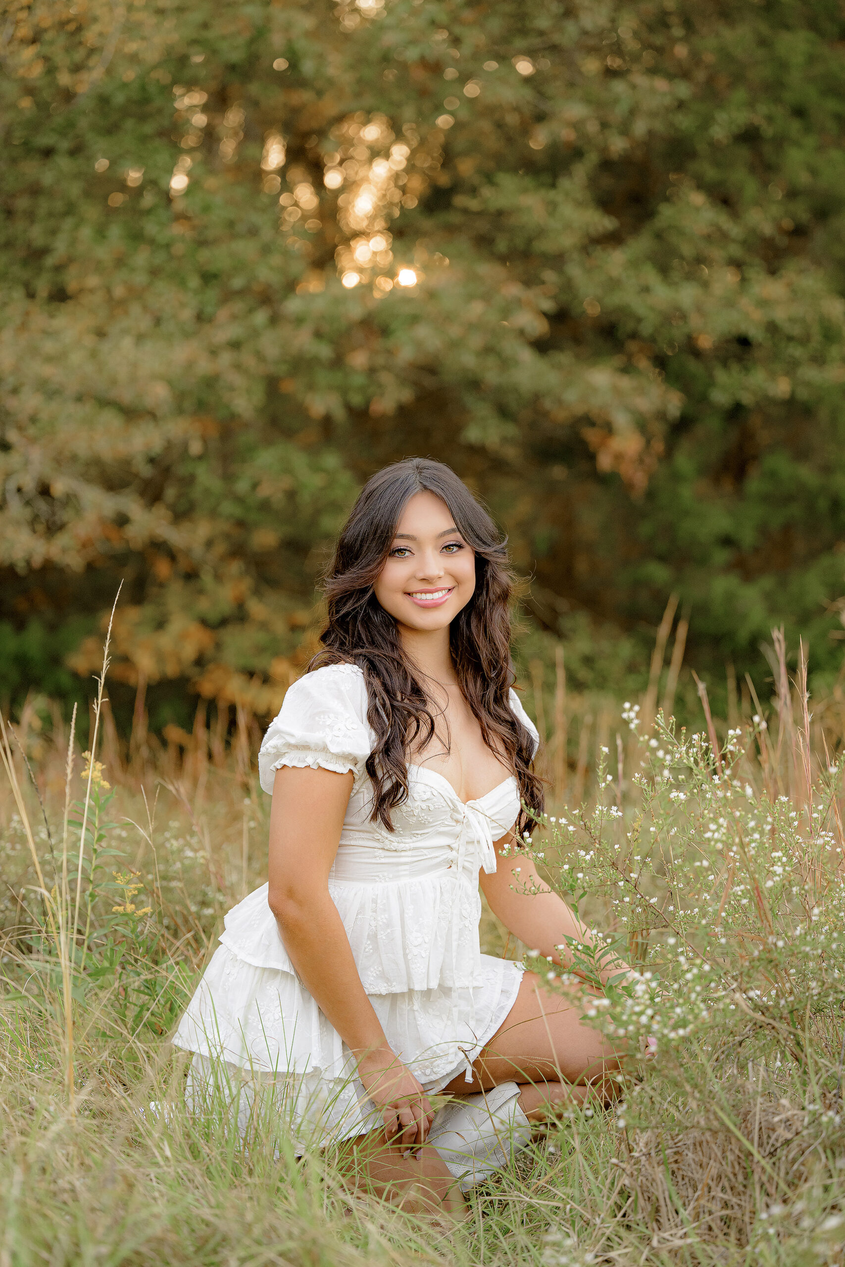 Dark haired class of 2025 grad girl sits playfully in the grass, wearing a white dress and cowboy boots