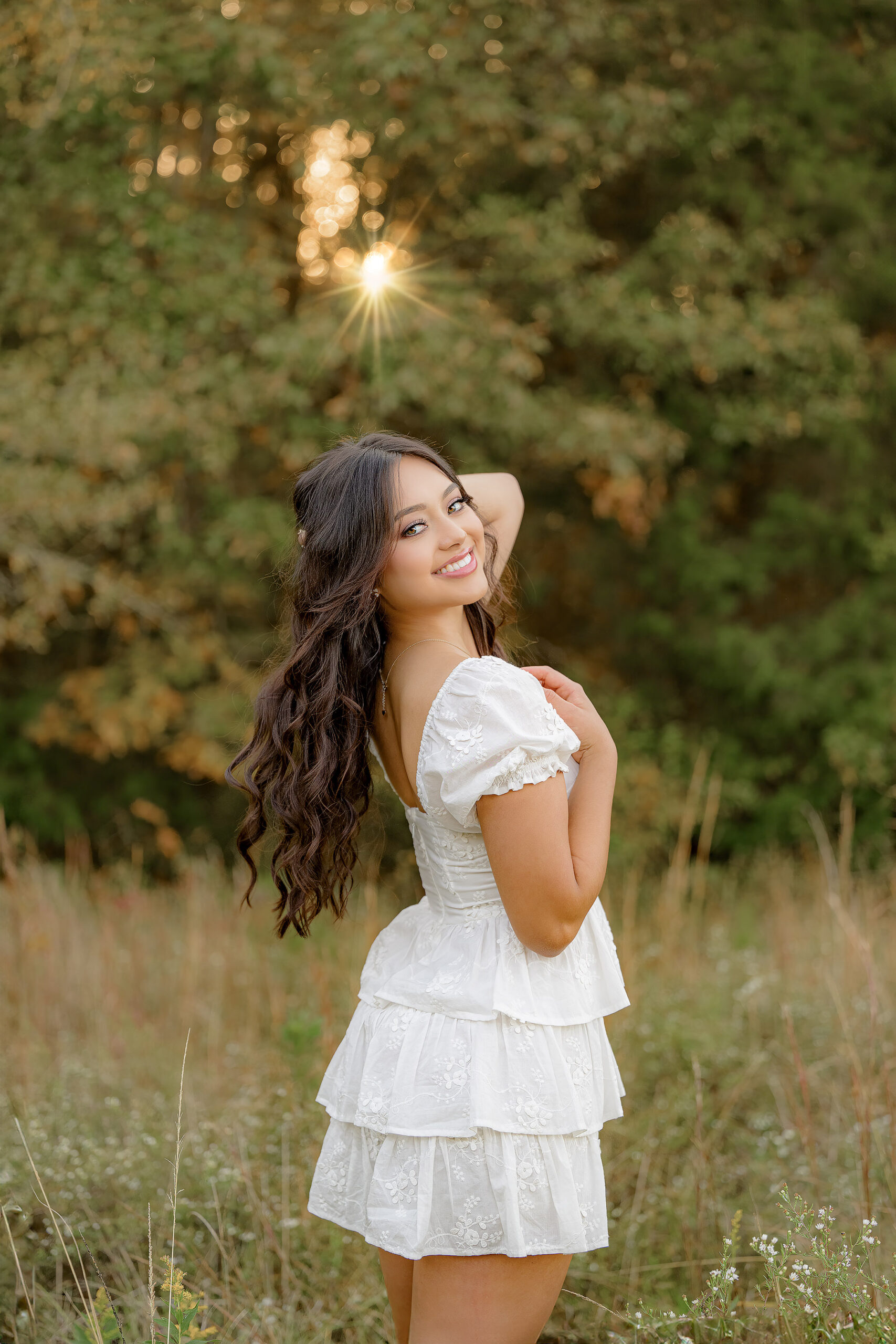 grad has her hand in her hair looking over her shoulder, dressed in a white dress standing in tall grass