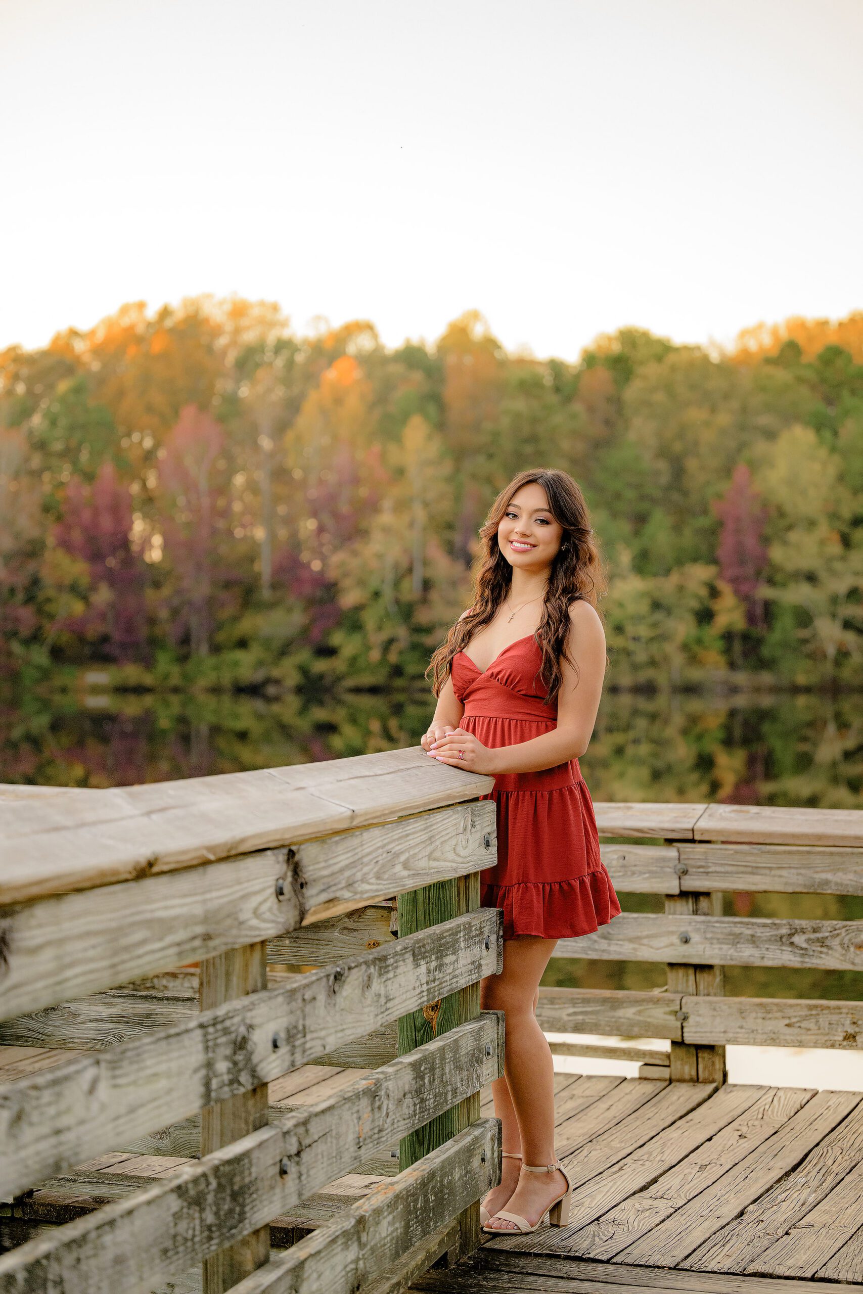 A Lakeside image with a dark haired, senior girl, dressed in a red dress, surrounded by fall colors