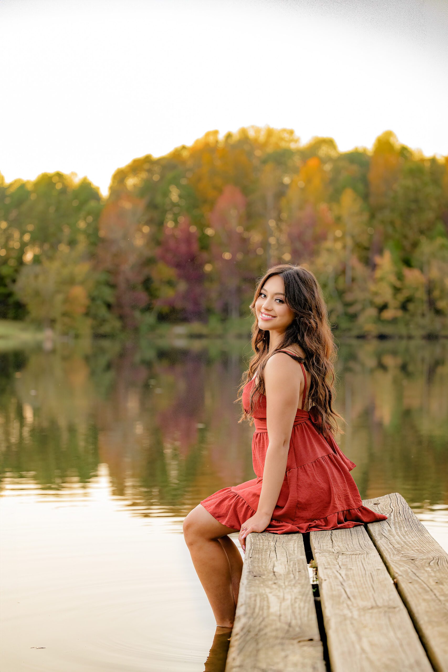 Class of 2025 Senior sits on the edge of a dock with her feet in the water surrounded by fall colors in a red dress