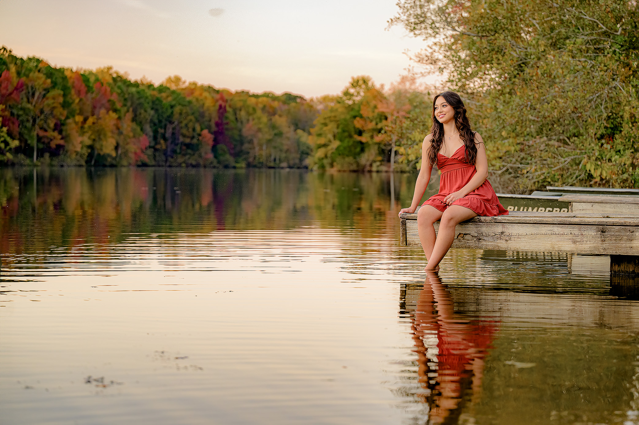 Senior girl looks off in the distance with her hair, flowing down around her, dressed in a red dress with her feet in the water and her reflection shining through surrounded by fall colors