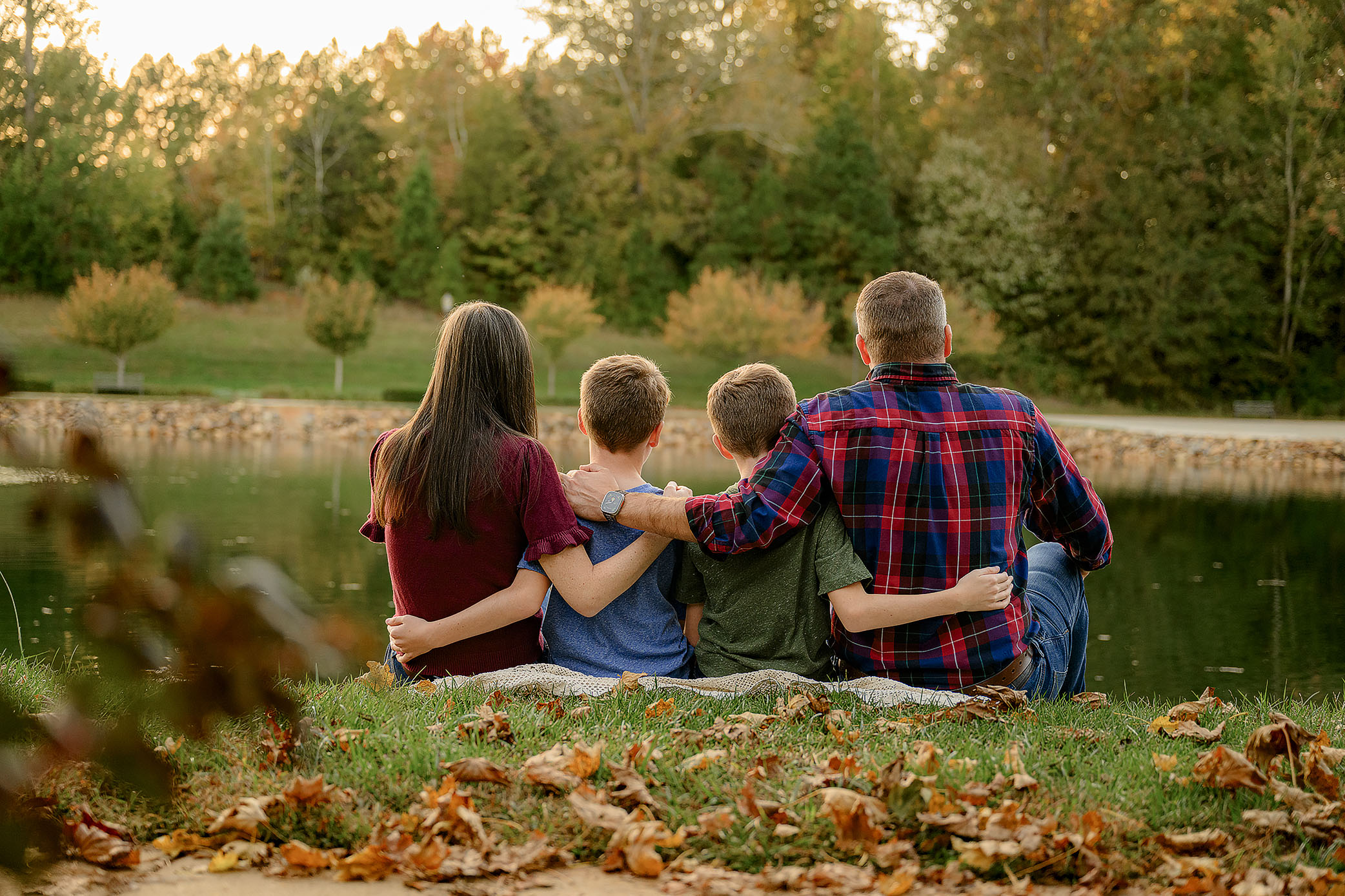 family sits on blanket facing away from the camera looking toward a pond