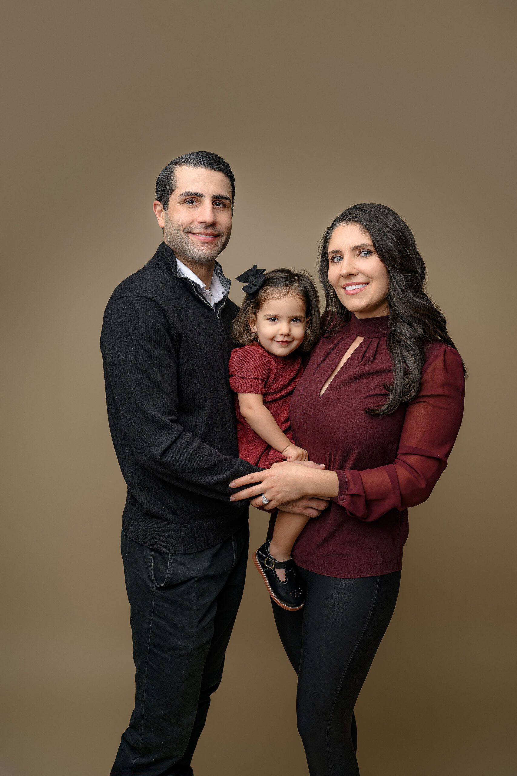 Mom dressed in red top and black pants with dad dressed in black holding their two year old daughter wearing a red dress, black shoes, and black hair bow