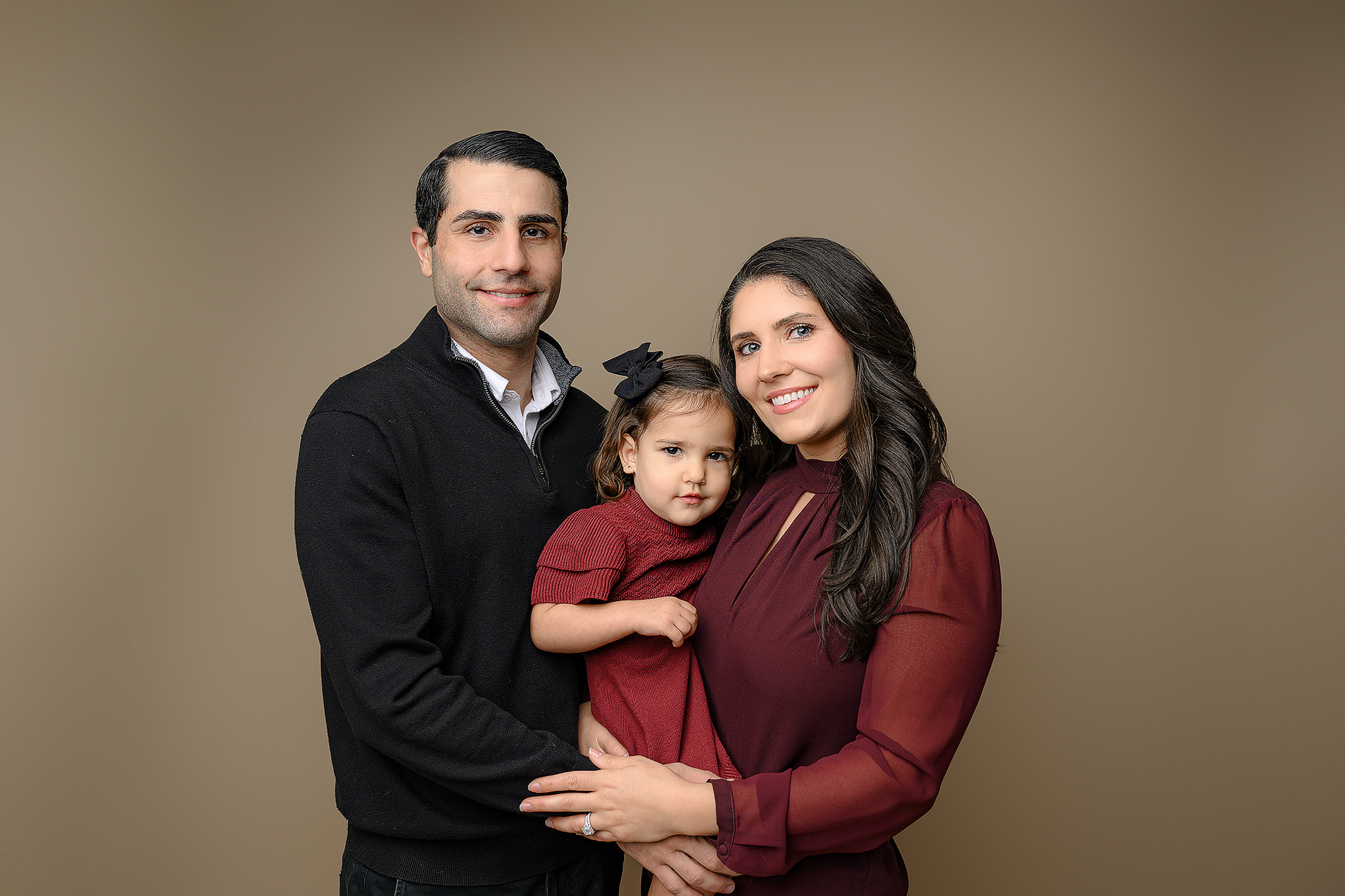 Mom dressed in red top and black pants with dad dressed in black holding their two year old daughter wearing a red dress, black shoes, and black hair bow in Milestone two year old photo session