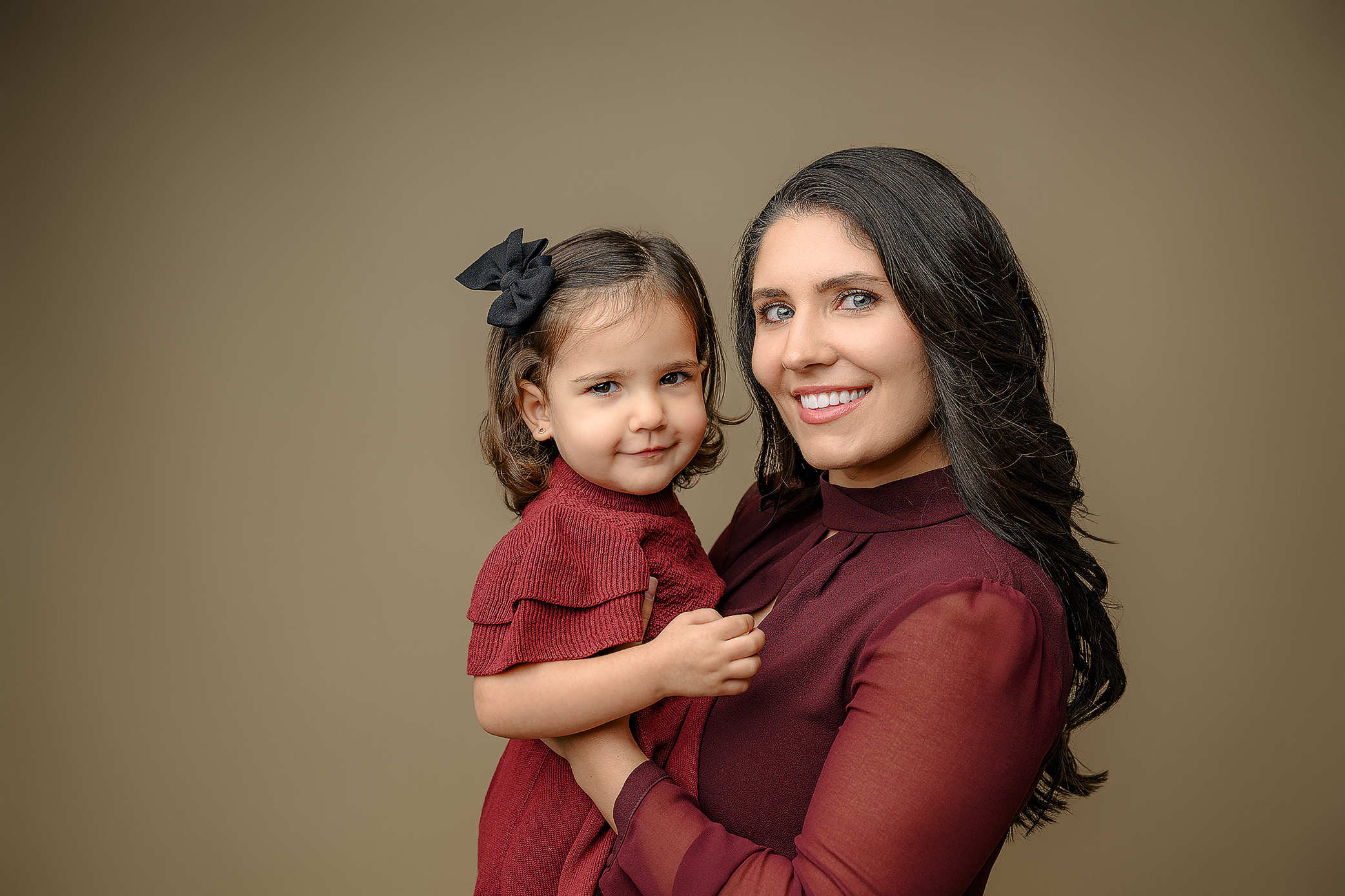 Mom dressed in red top and black holding her two year old daughter wearing a red dress and black hair bow