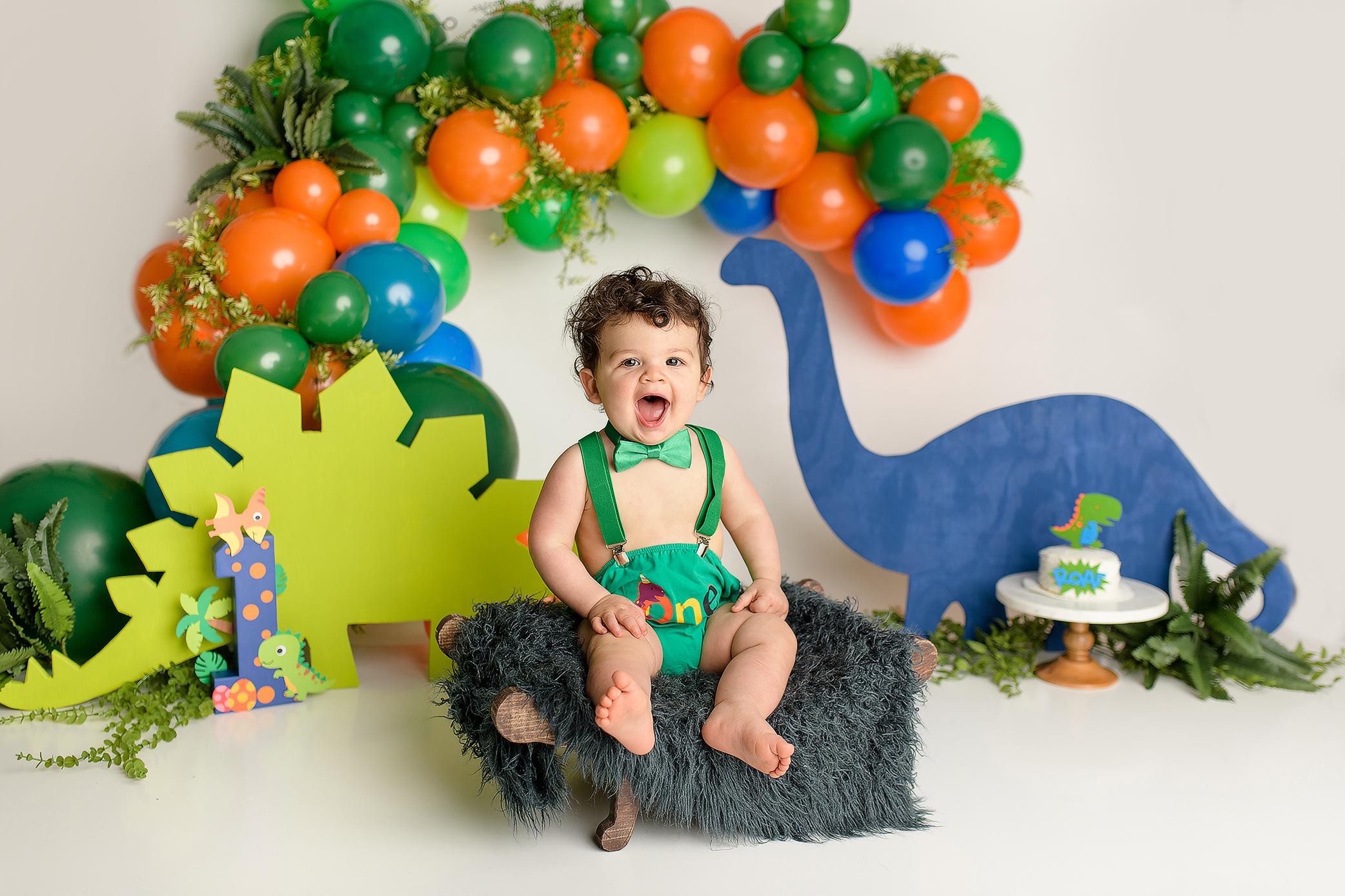 A one-year-old boy sitting in a colorful dinosaur-themed cake smash setup. He is dressed in a green diaper cover, green suspenders, and a green bow tie. The background features vibrant orange, green, and blue dinosaur cutouts, creating a playful atmosphere as the little boy smiles, enjoying his surroundings.