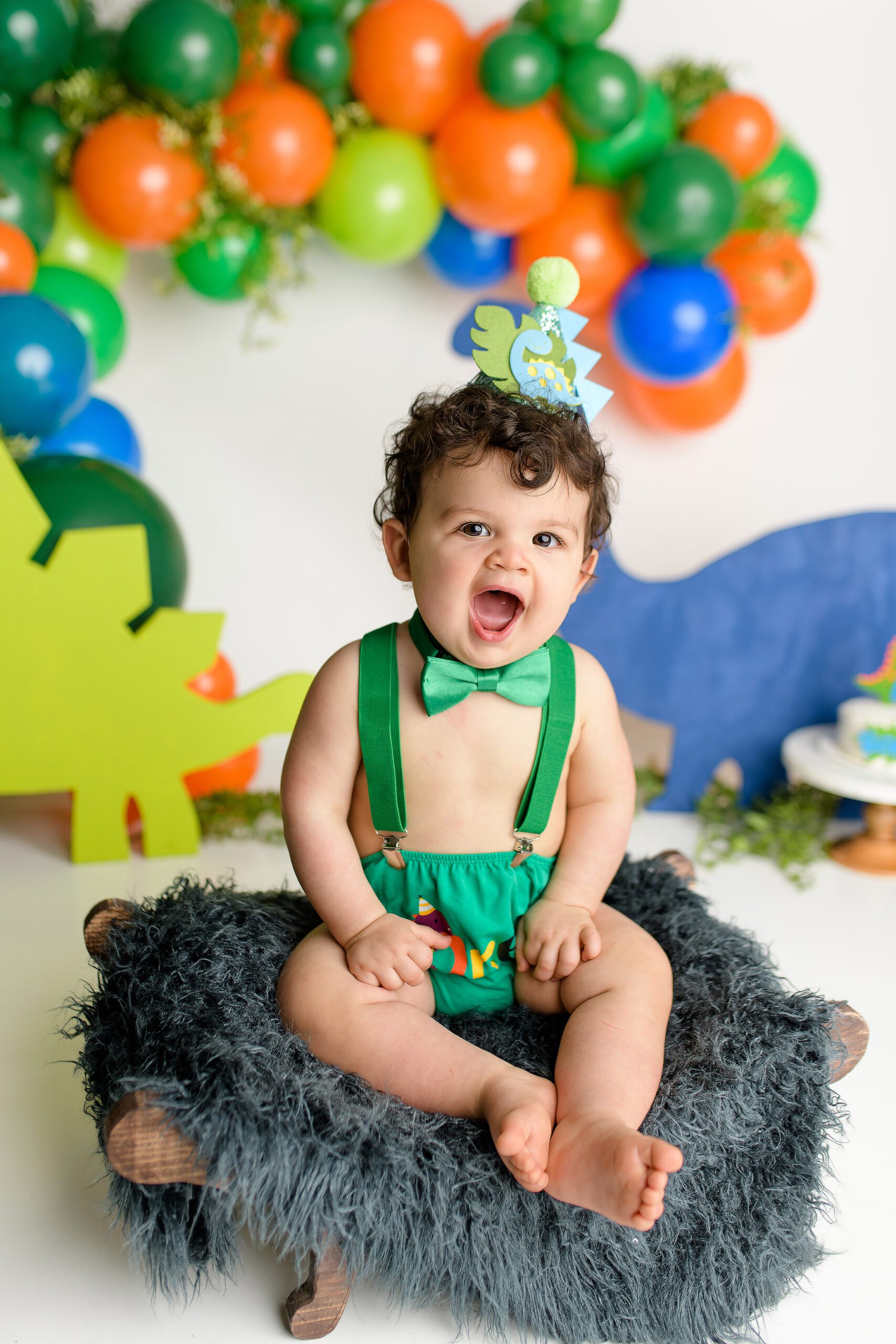 A one-year-old boy dressed in a green diaper cover, green suspenders, and a green bow tie, surrounded by orange, green, and blue dinosaur decorations. He sits in his cake smash setup with a dinosaur hat on his head, smiling as he enjoys his special moment.