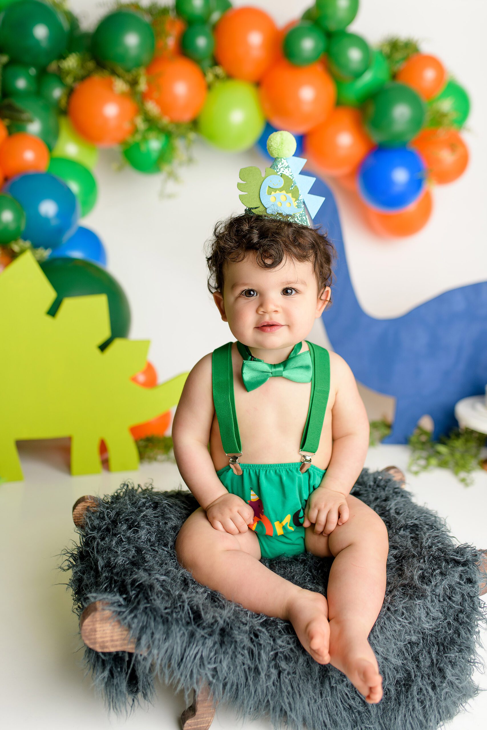 A one-year-old boy in a playful pose at his dinosaur-themed cake smash setup. He’s wearing a green diaper cover, green suspenders, and a green bow tie, surrounded by vibrant orange, green, and blue dinosaur cutouts. A dinosaur hat sits on his head as he looks curiously at the photographer.