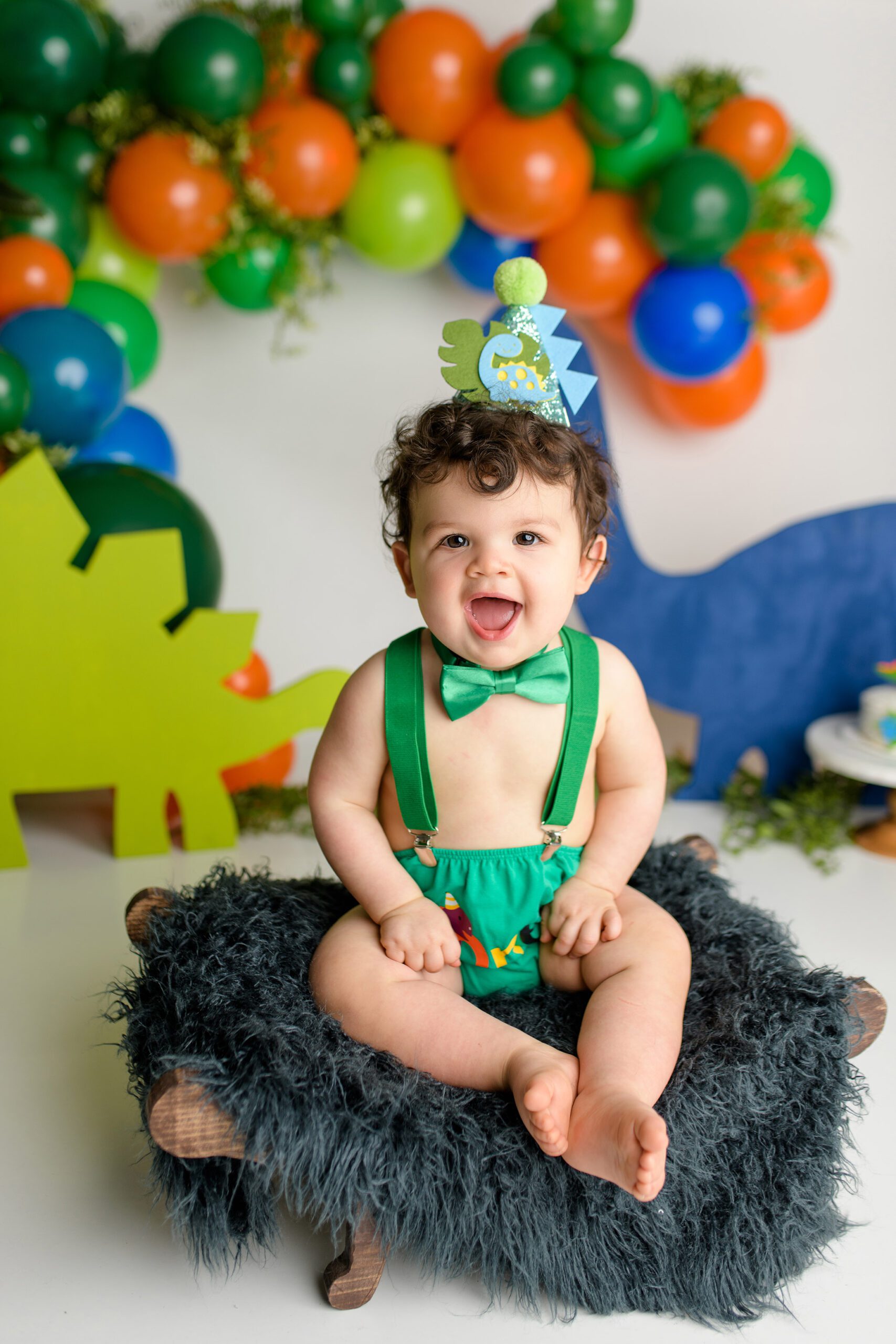 A joyful one-year-old boy sitting in his dinosaur-themed cake smash scene. He is dressed in a green diaper cover, green suspenders, and a green bow tie. Bright orange, green, and blue dinosaur cutouts surround him, and a fun dinosaur hat sits on his head as he looks around with excitement.