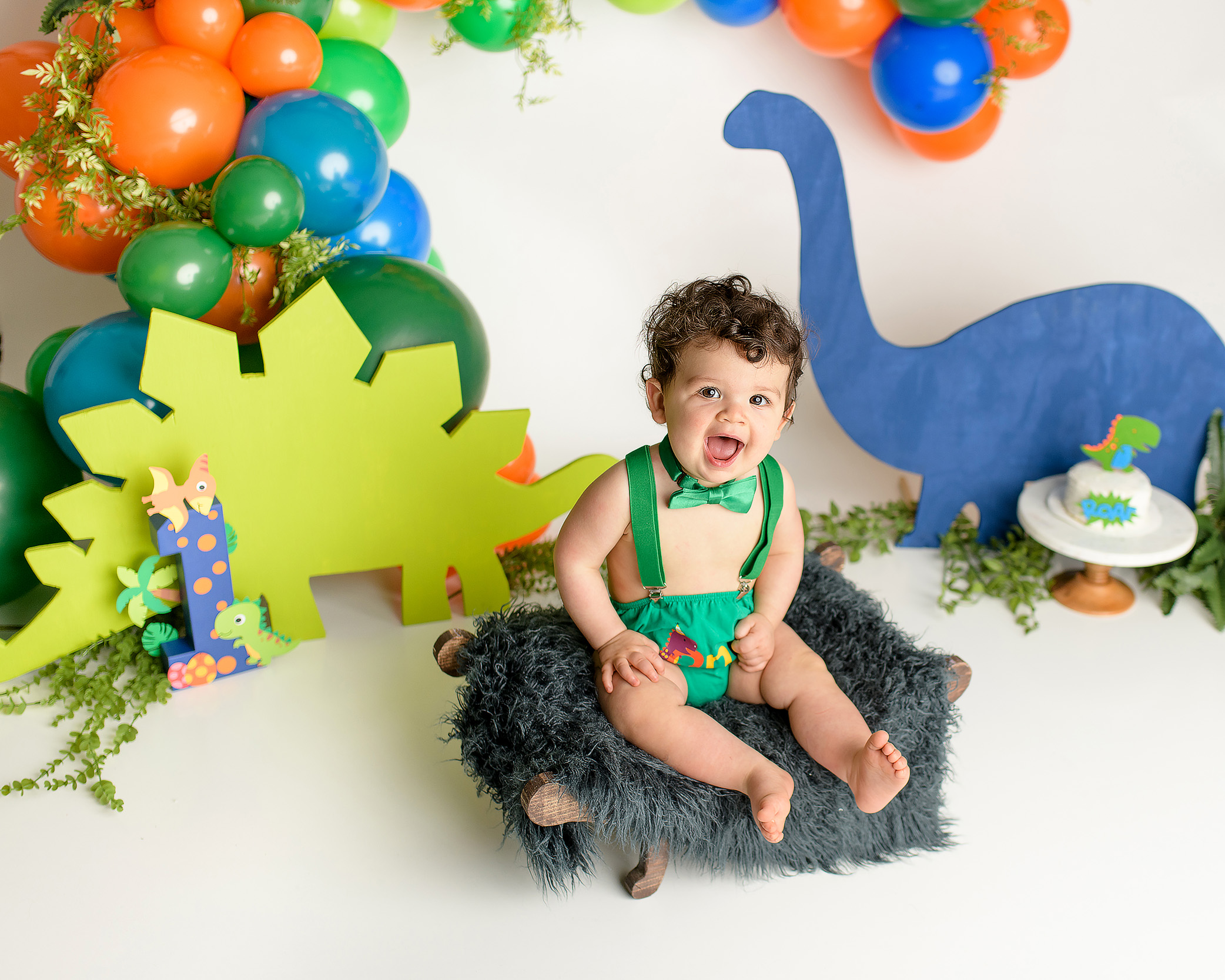 A one-year-old boy sitting in his one year old dinosaur-themed cake smash setup, looking curiously at the cake in front of him. He’s dressed in a green diaper cover, green suspenders, and a green bow tie. The backdrop features orange, green, and blue dinosaur cutouts as he has a playful good time before his smash.