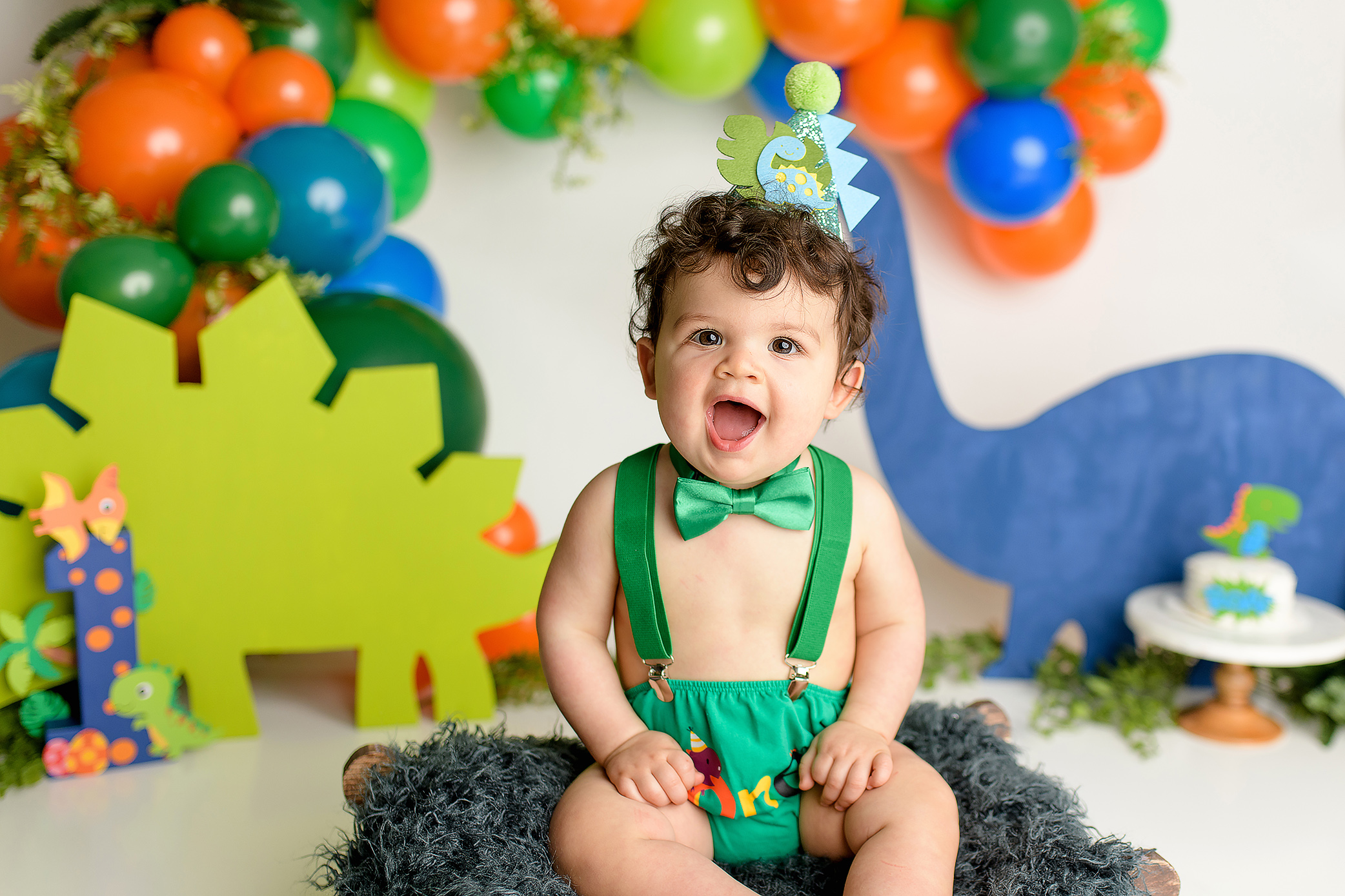 A one-year-old boy sitting happily in a dinosaur-themed cake smash setup, wearing a green diaper cover, suspenders, and bow tie. The backdrop features colorful orange, green, and blue dinosaur decorations. He’s wearing a cute dinosaur hat and enjoying the fun atmosphere.