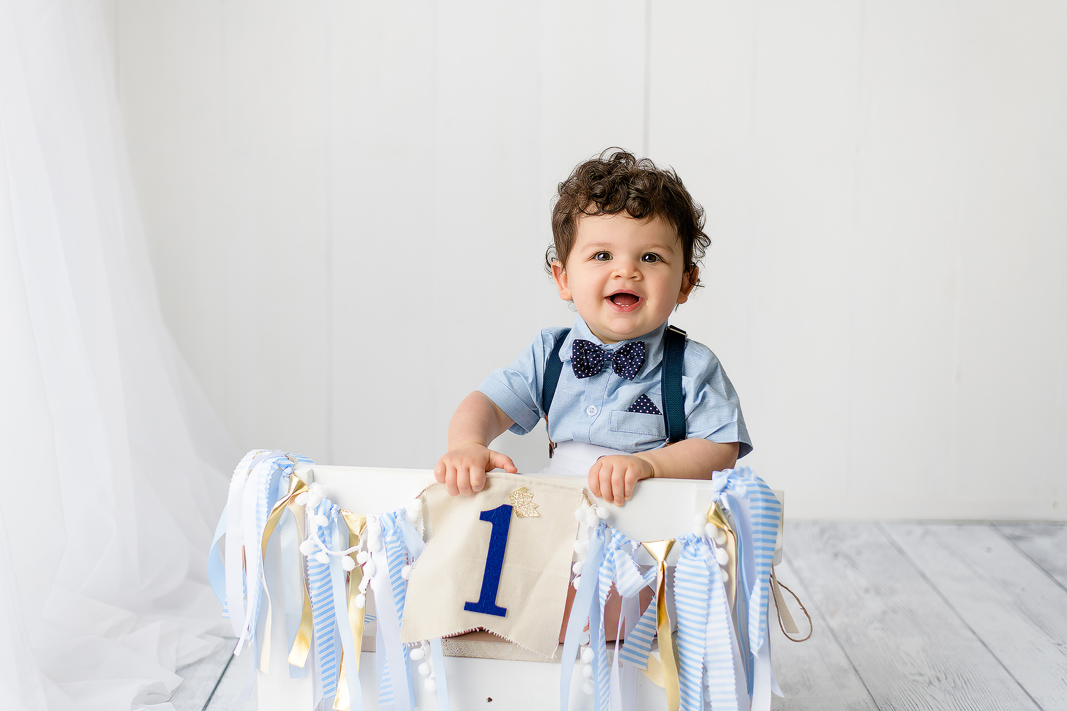 Stylish birthday boy sitting on a white bench, dressed in a navy bow tie, blue shirt, and white shorts for his first milestone photos.