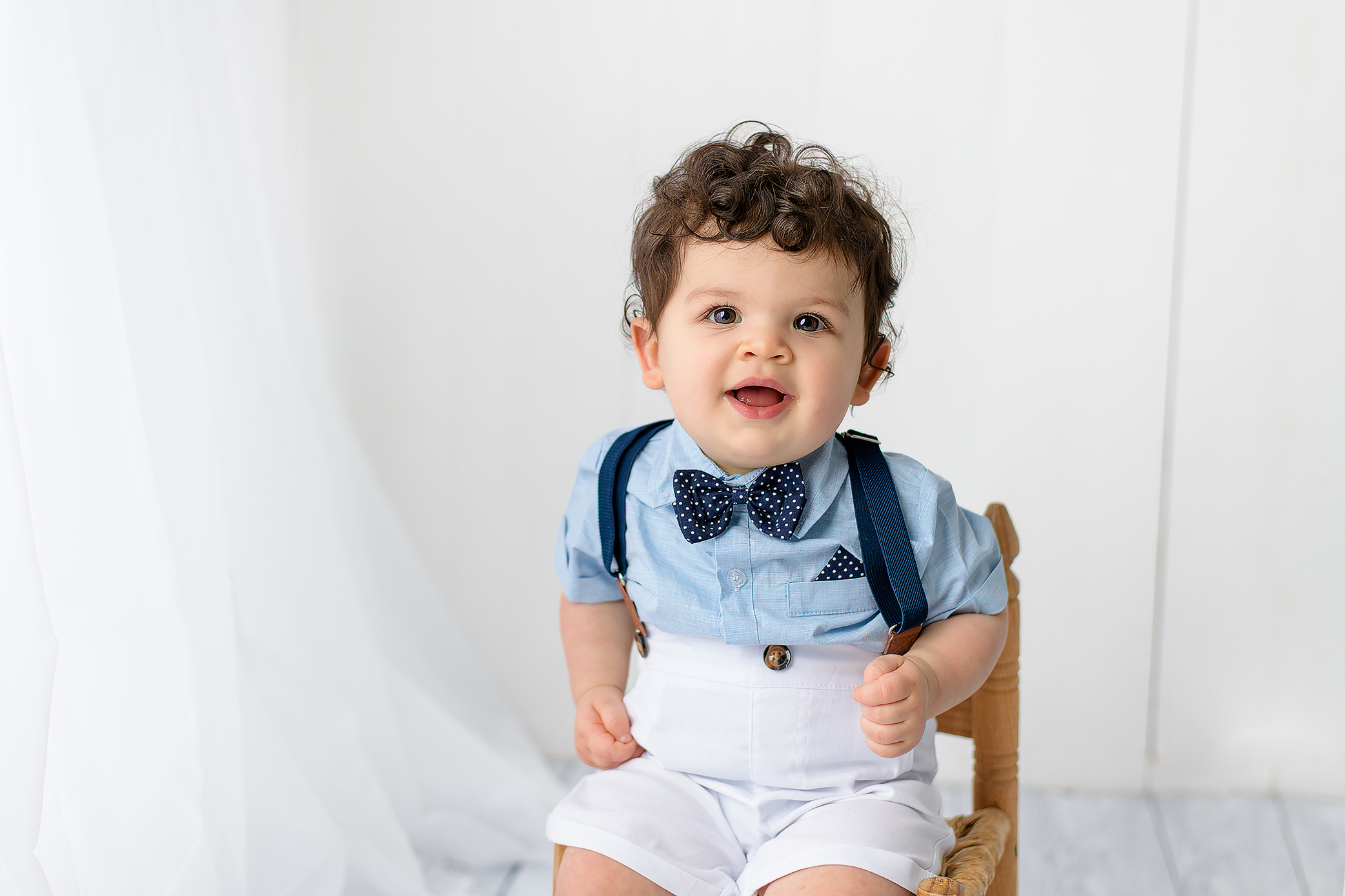 A one-year-old boy sitting on a colorful dinosaur-themed cake smash setup. He’s wearing a green diaper cover, green suspenders, and a green bow tie. The backdrop features orange, green, and blue dinosaur cutouts. The little boy smiles with a playful dinosaur hat resting on his head.