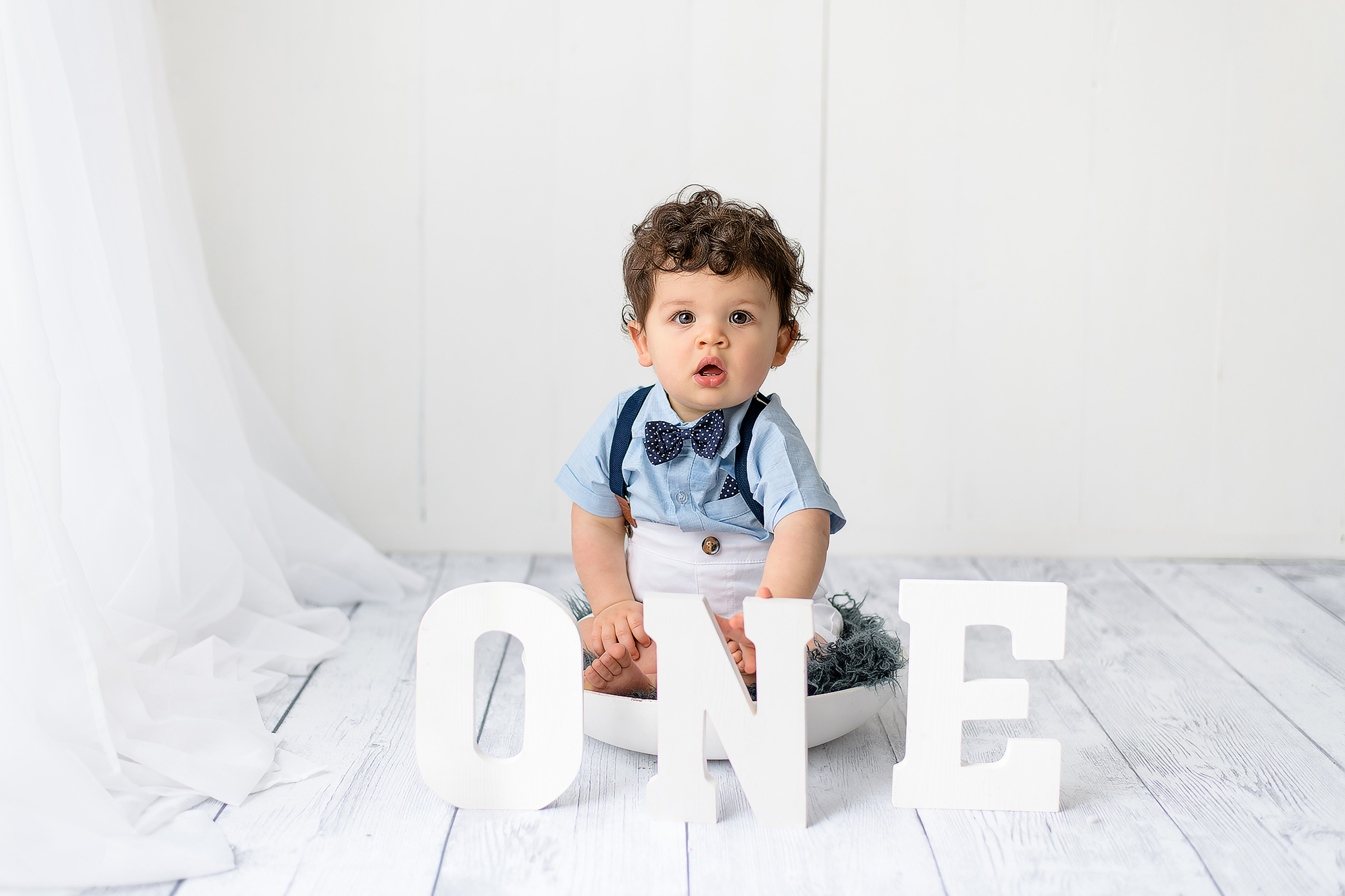 Milestone photos of a sweet one-year-old boy with dark hair, dressed in a classic outfit and sitting in a bowl with ONE letters in front of him in a classic white set up