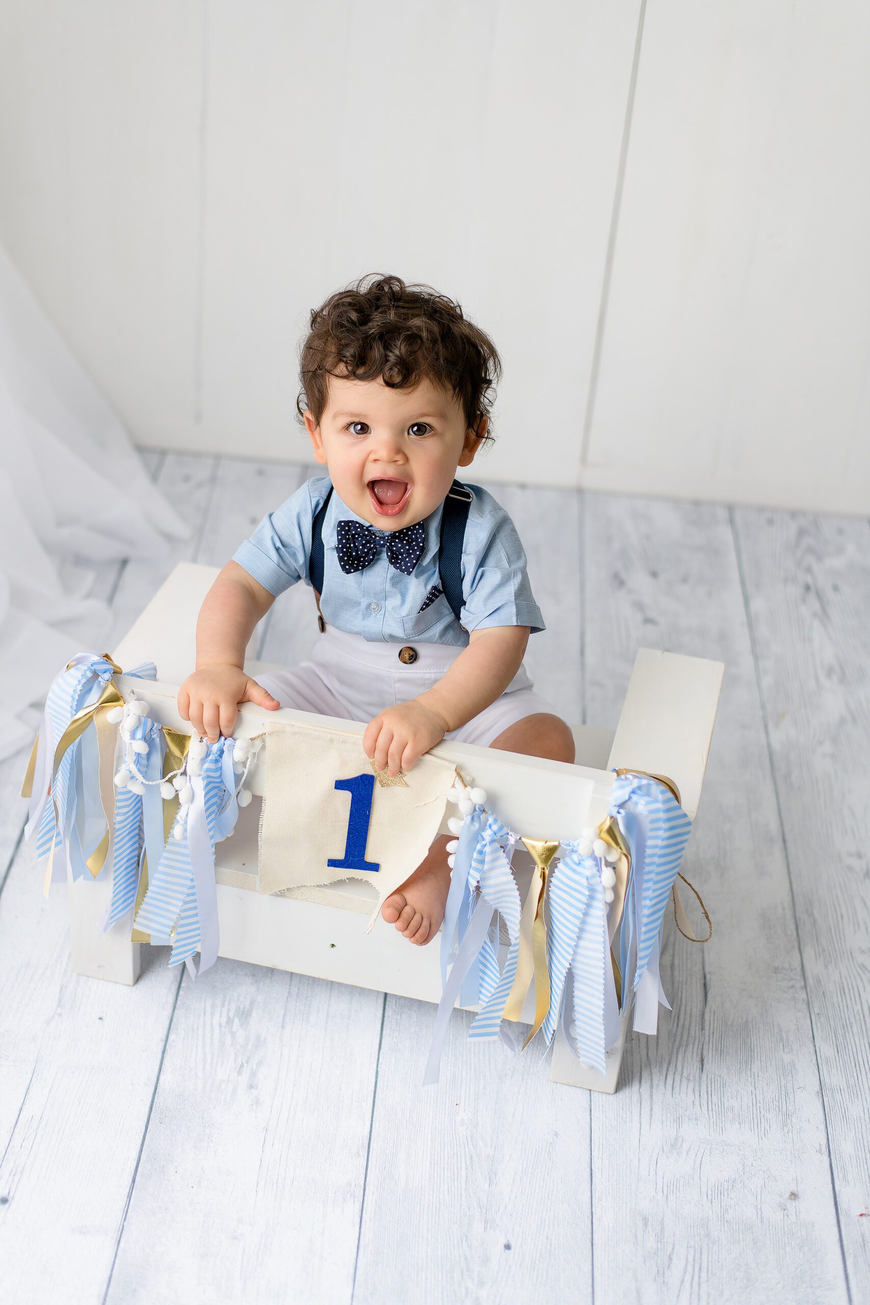 A one-year-old boy with dark hair sits on a small white bench wearing a blue shirt, navy bow tie, and white shorts, smiling sweetly during his one year old birthday photoshoot.