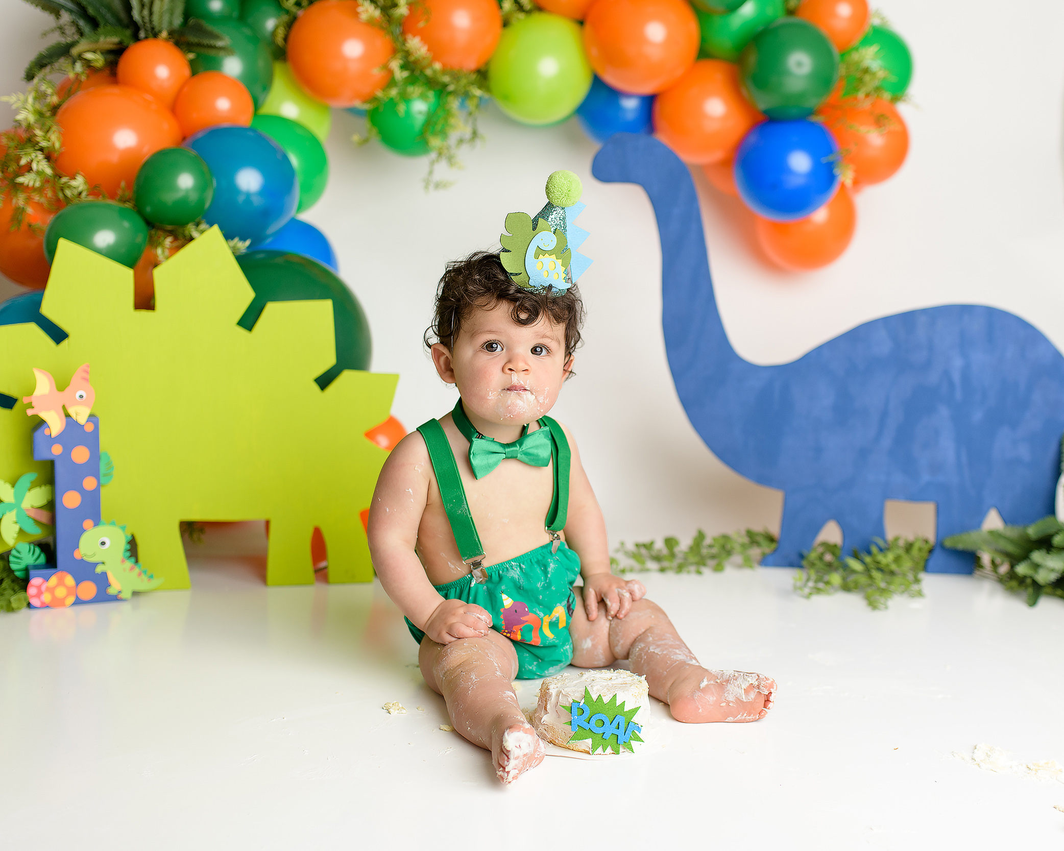 A one-year-old boy tasting the cake during his dinosaur-themed cake smash. He’s wearing a green diaper cover, green suspenders, and a green bow tie. The cake is in front of him as he reaches out with his hands to take a bite, surrounded by colorful orange, green, and blue dinosaur cutouts.