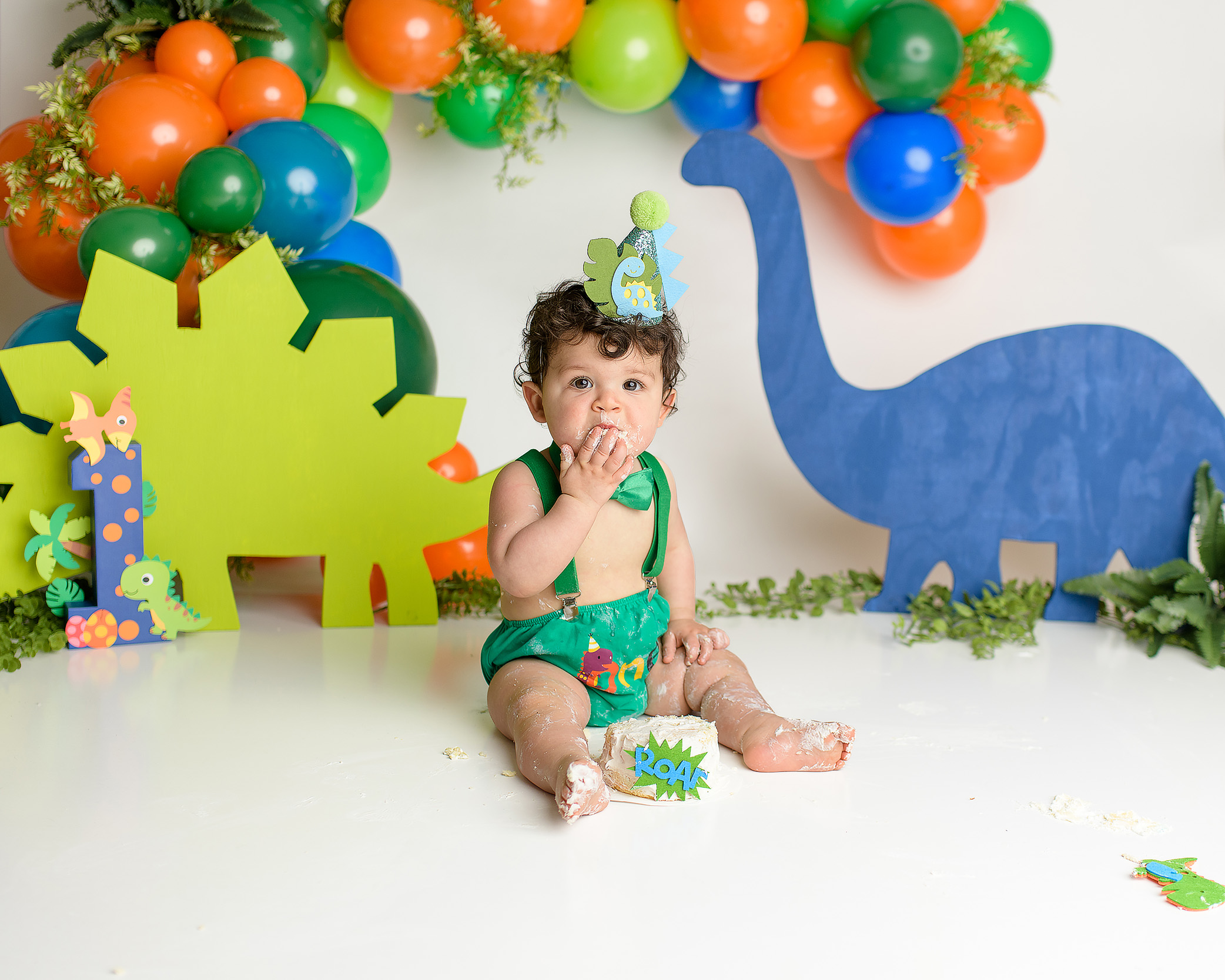A one-year-old boy laughing joyfully as he smashes the cake in his dinosaur-themed cake smash setup. He’s dressed in a green diaper cover, green suspenders, and a green bow tie. The backdrop is decorated with orange, green, and blue dinosaur cutouts, as he enjoys the playful mess of the cake and giggles in delight.