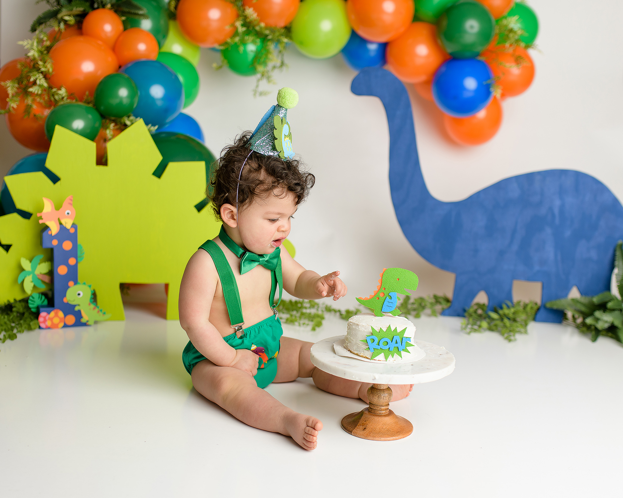 A one-year-old boy sitting in his dinosaur-themed cake smash setup, looking curiously at the cake in front of him. He’s dressed in a green diaper cover, green suspenders, and a green bow tie. The backdrop features orange, green, and blue dinosaur cutouts as he focuses on the cake with a curious expression.