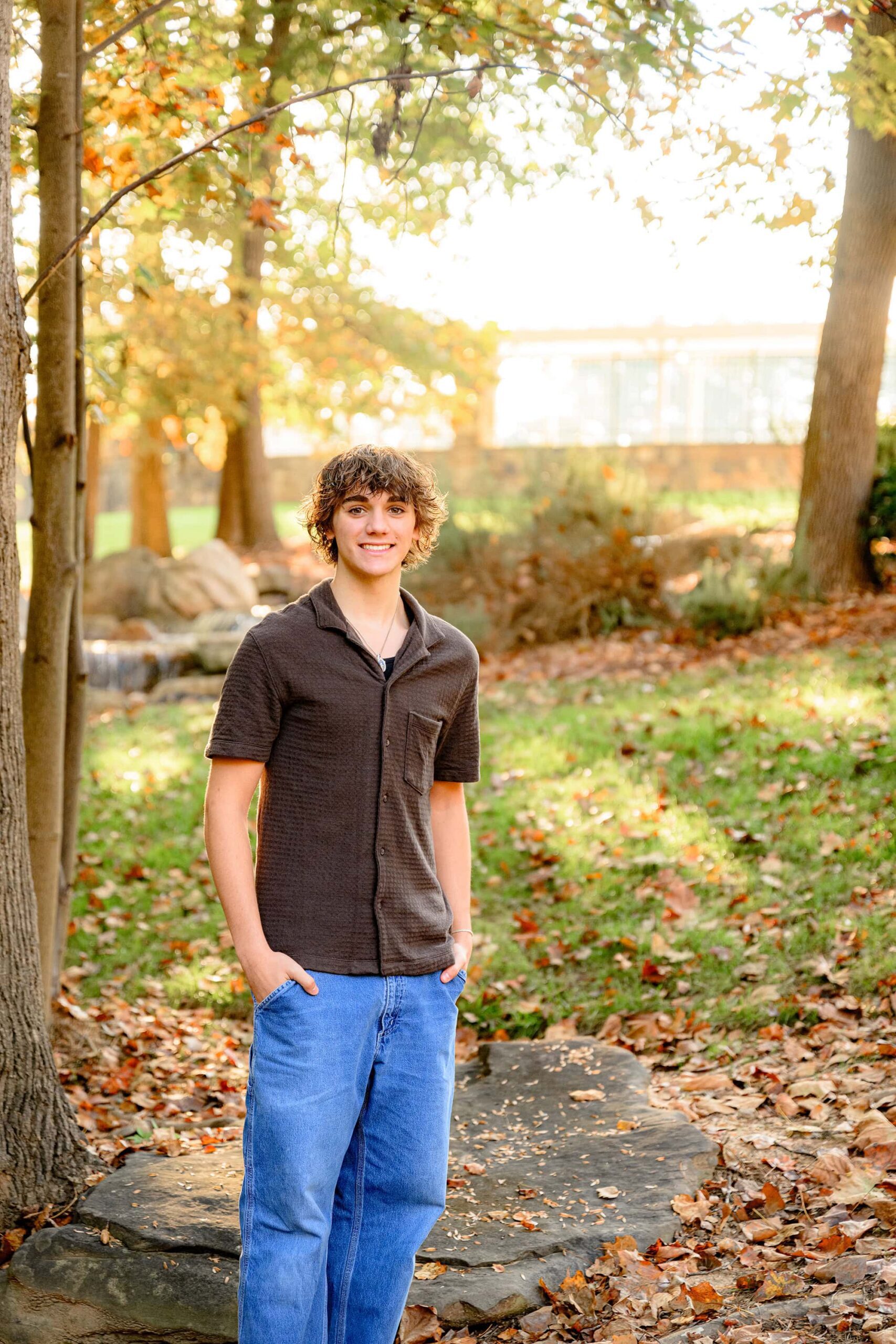 dark hair senior boy standing wearing jeans and a brown shirt with his hands in his pockets during his waxhaw nc senior photo session