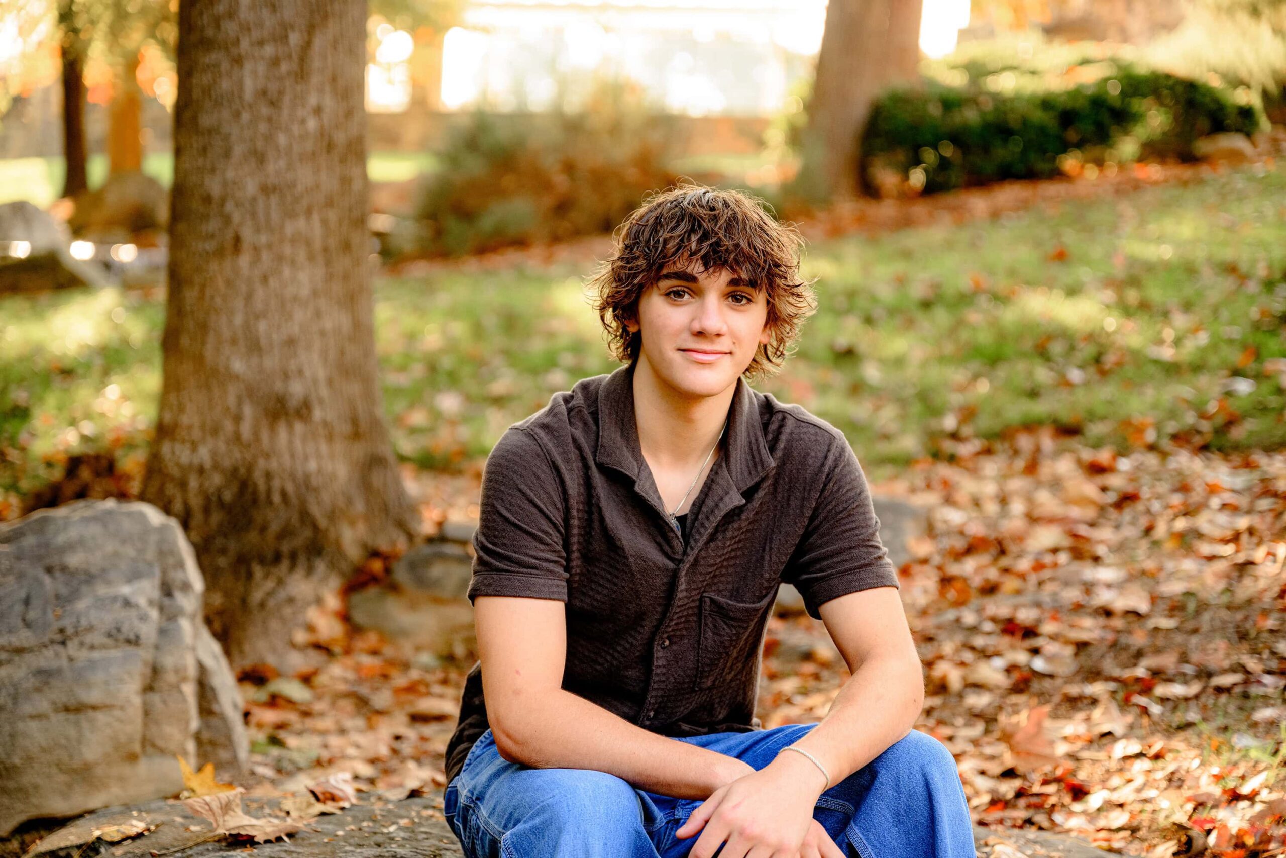 class of 2025 senior boy with dark shaggy hair sitting on rock during his senior session surrounded by fall colors and morning golden light