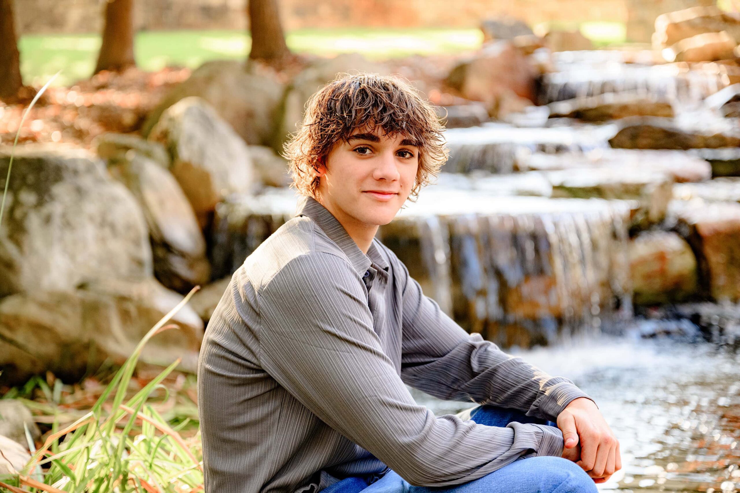 sitting on a rock dressed in grey the senior boy is looking toward the camera as the golden light shines on the back of his hair