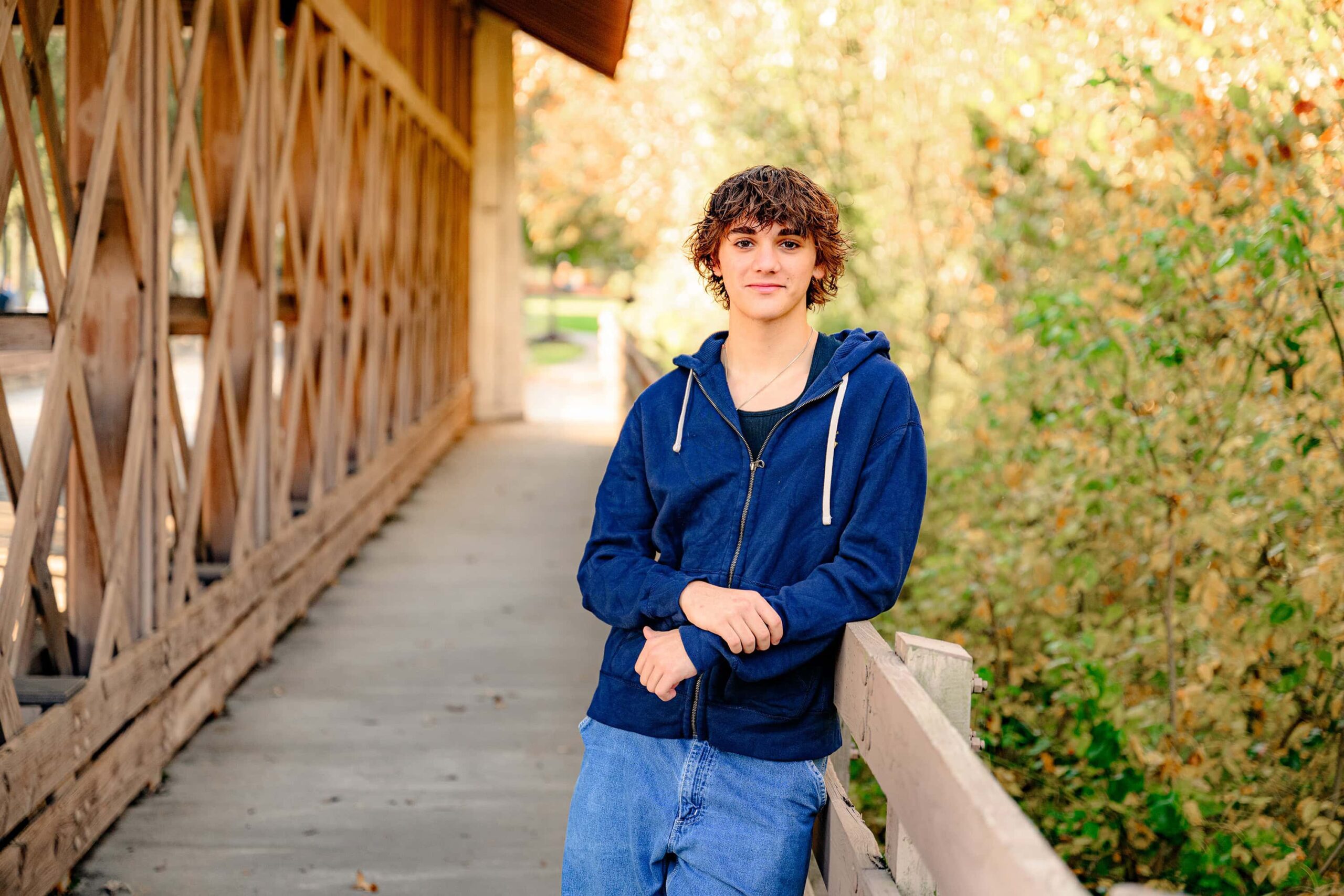 Waxhaw, NC Class of 2025 senior boy with shaggy dark hair leaning against a railing dressed in a blue jacket and jeans with the fall trees in perfect color and in bright golden light surrounding him