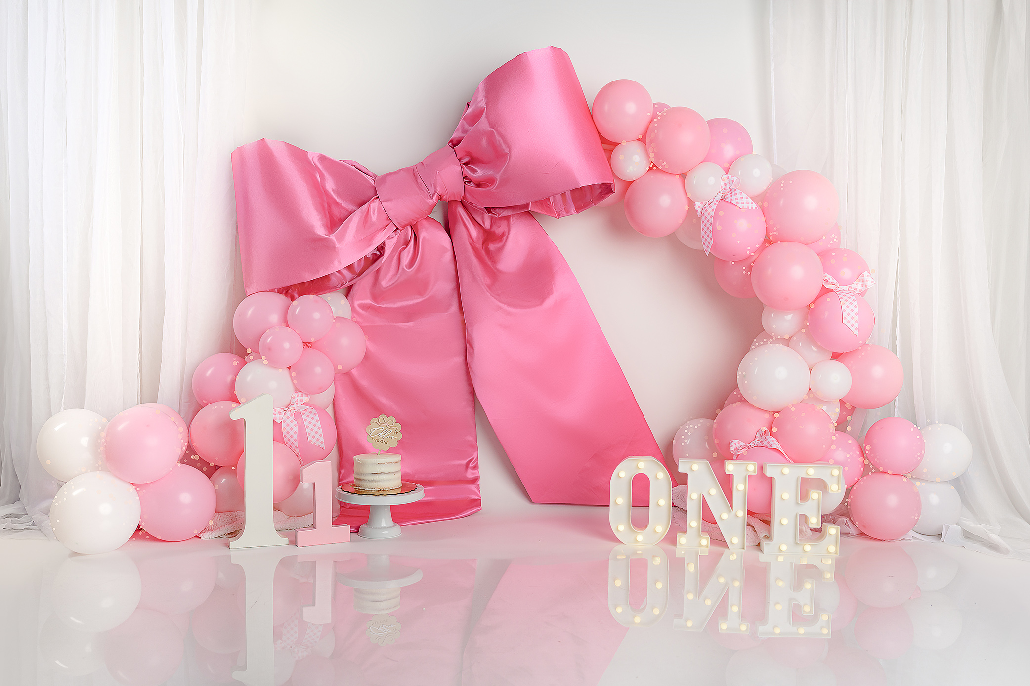 A giant pink satin bow steals the show in this baby milestone smash set complete with marquee letters, numbers, a cute cake and pink and white balloon arch