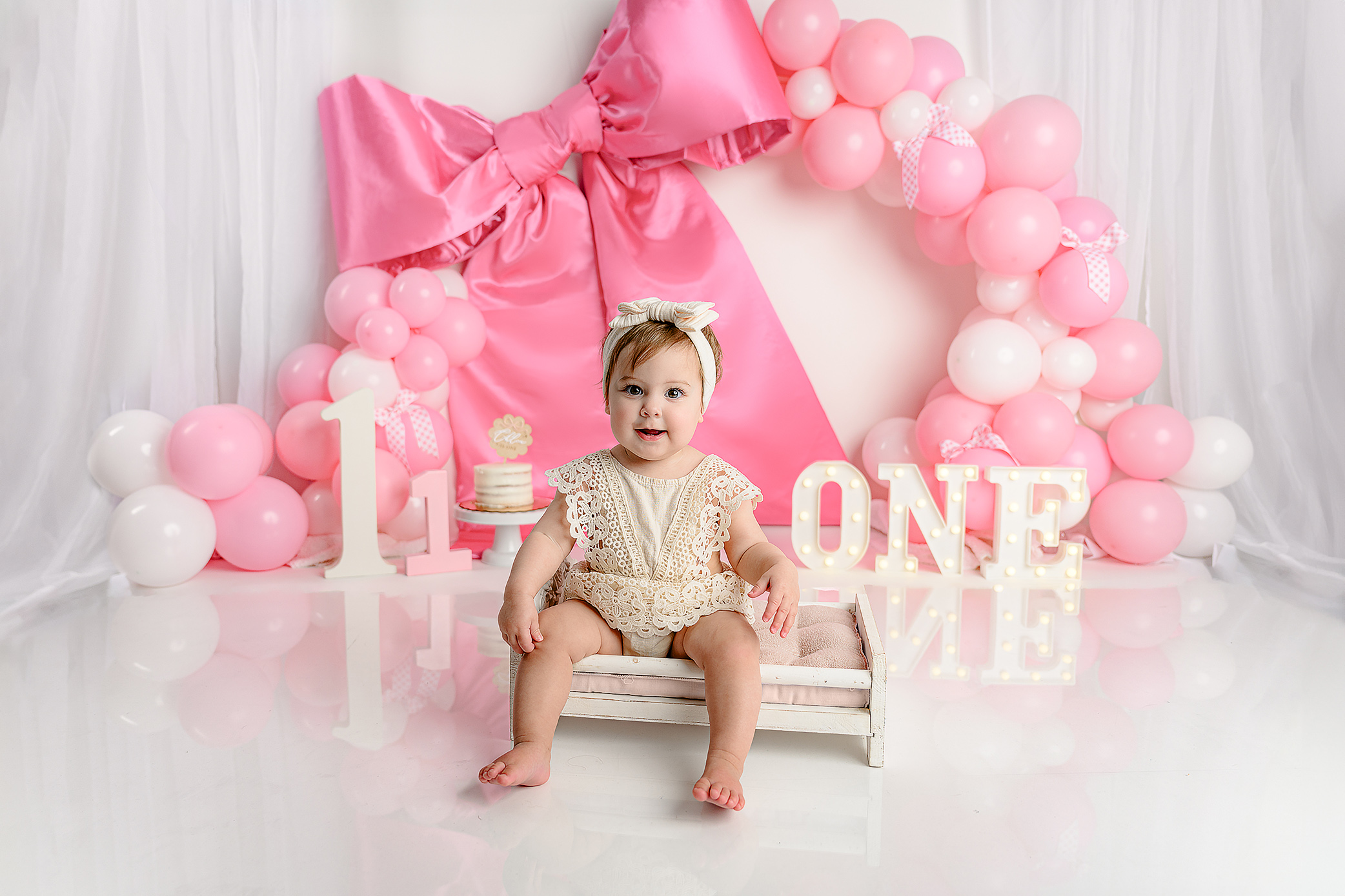 With dark hair, dressed in a cream outfit sitting in her cake smash set up that features a big satin, pink bow, pink and white balloons and fun features