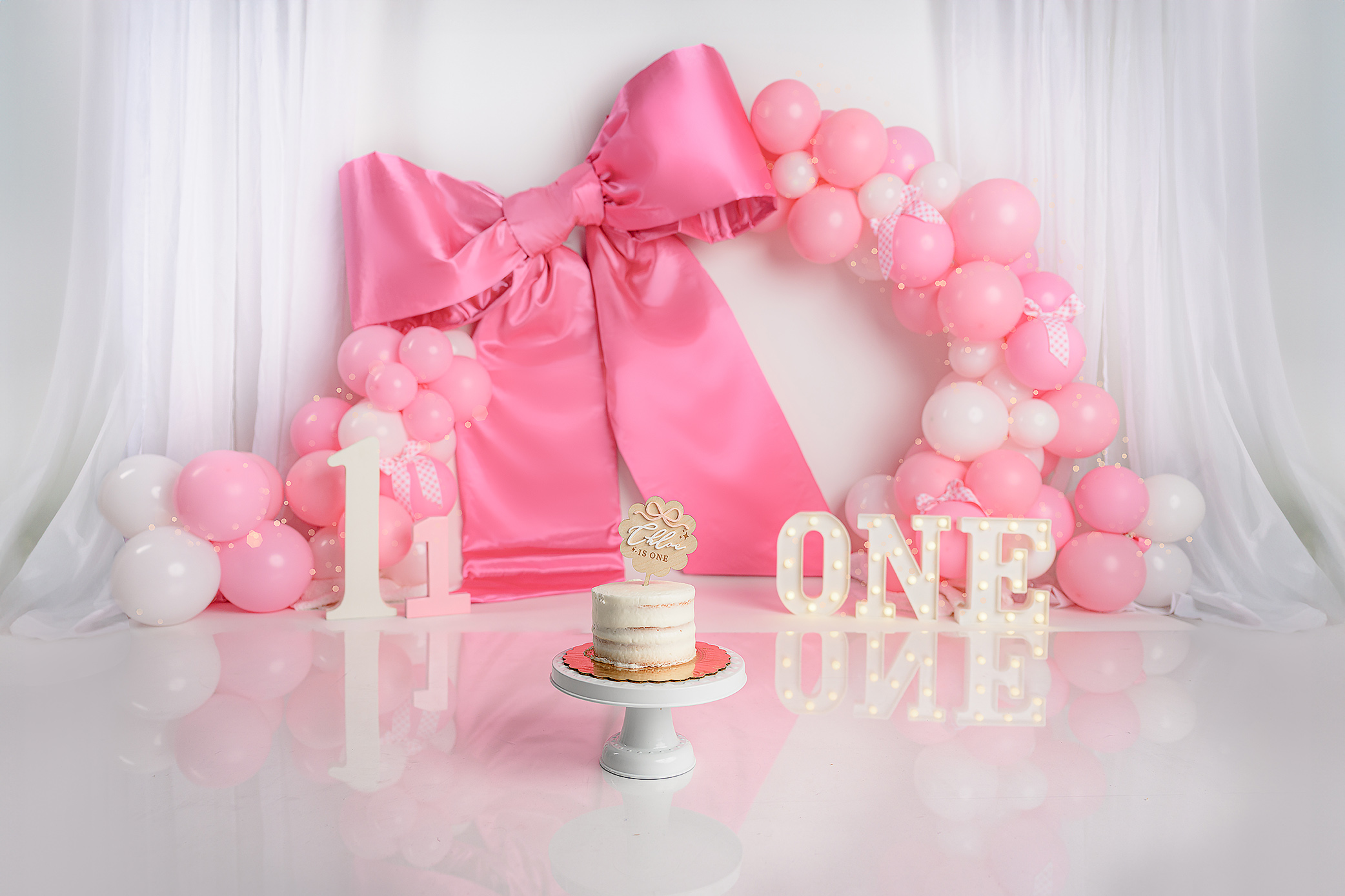 Pulled back photo of a cake smash set up for a little girl turning one that features a custom cake with a custom cake topper a giant satin bow, sheer curtains, framing, a balloon arch with pink and white balloons for a birthday session