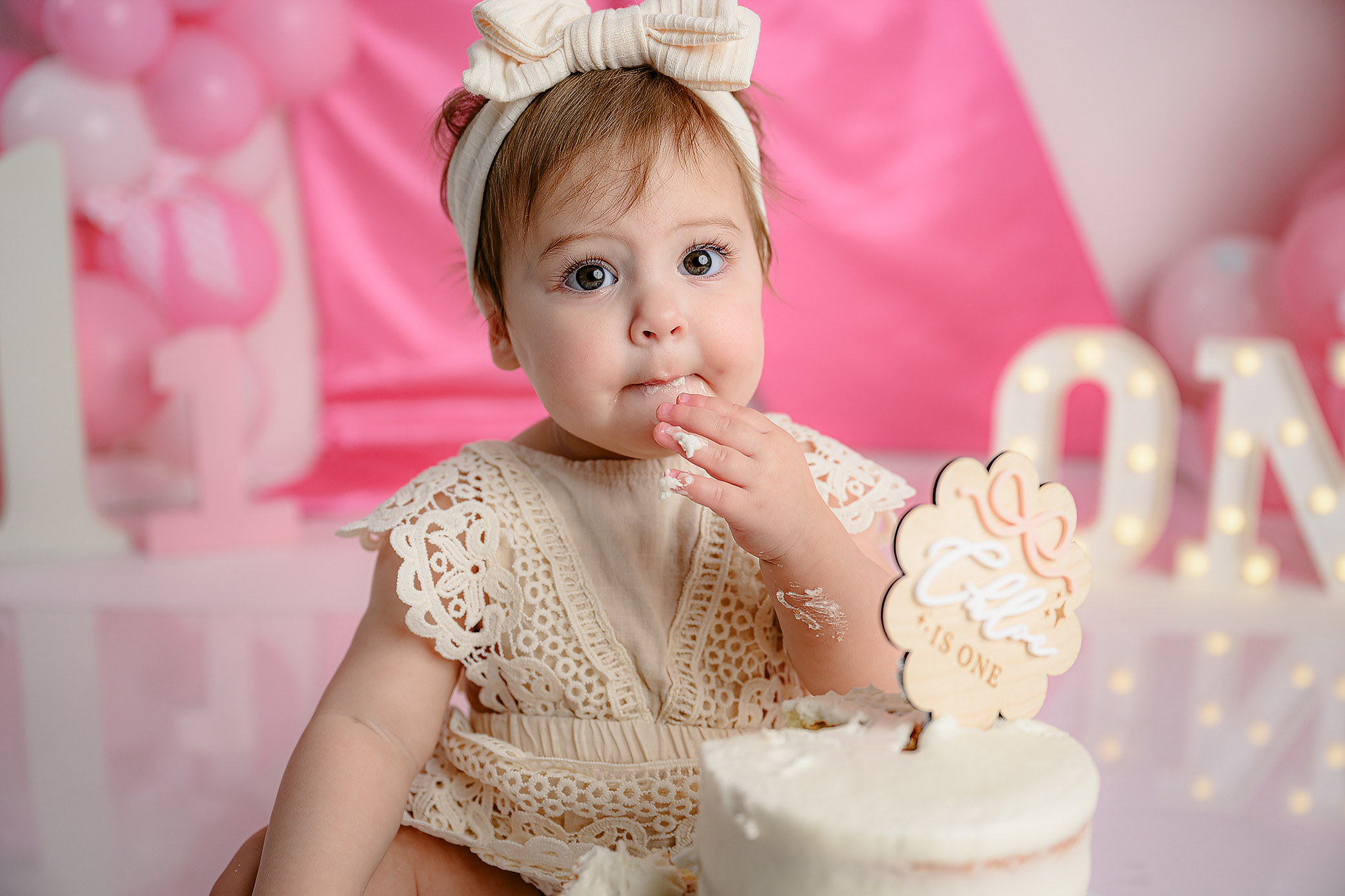 Close-up photo of a little girl dressed in cream with a bow on her head, eating a bite of cake during her milestone session in Waxhaw, North Carolina
