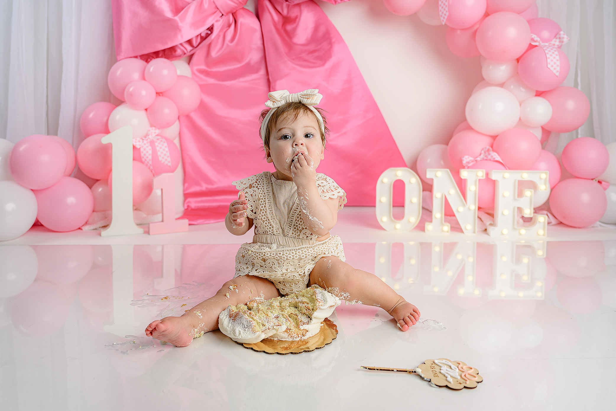 Little girl taking a big bite of her cake during her session that features marquee ONE letters, satin bow and pink and white balloons