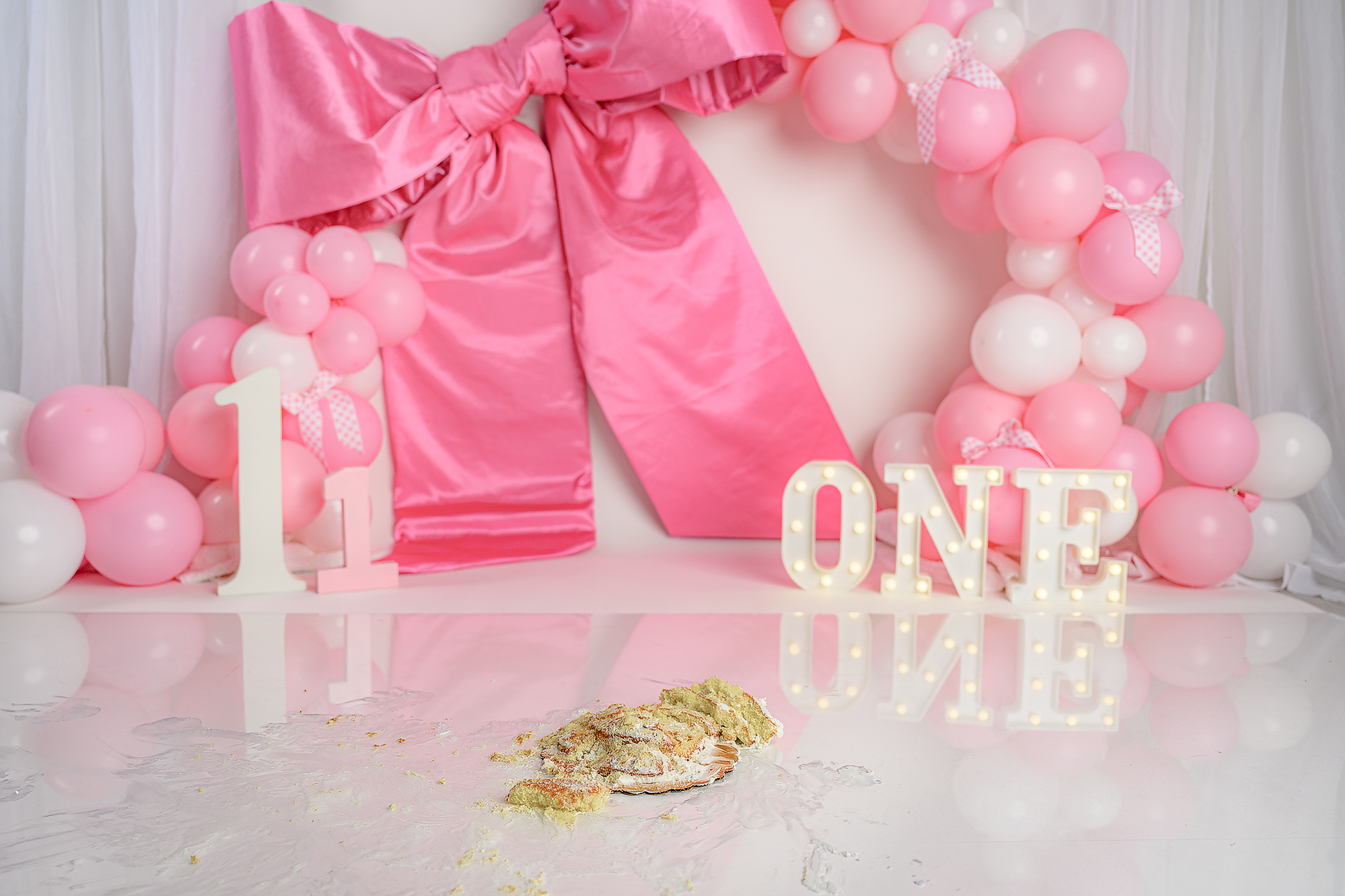 Final image from a cake smash session that shows the cake obliterated along with the super fun set up of a giant pink bow marquee ONE letters, the numbers, one pink and white balloons and white sheer curtains with gingham trim