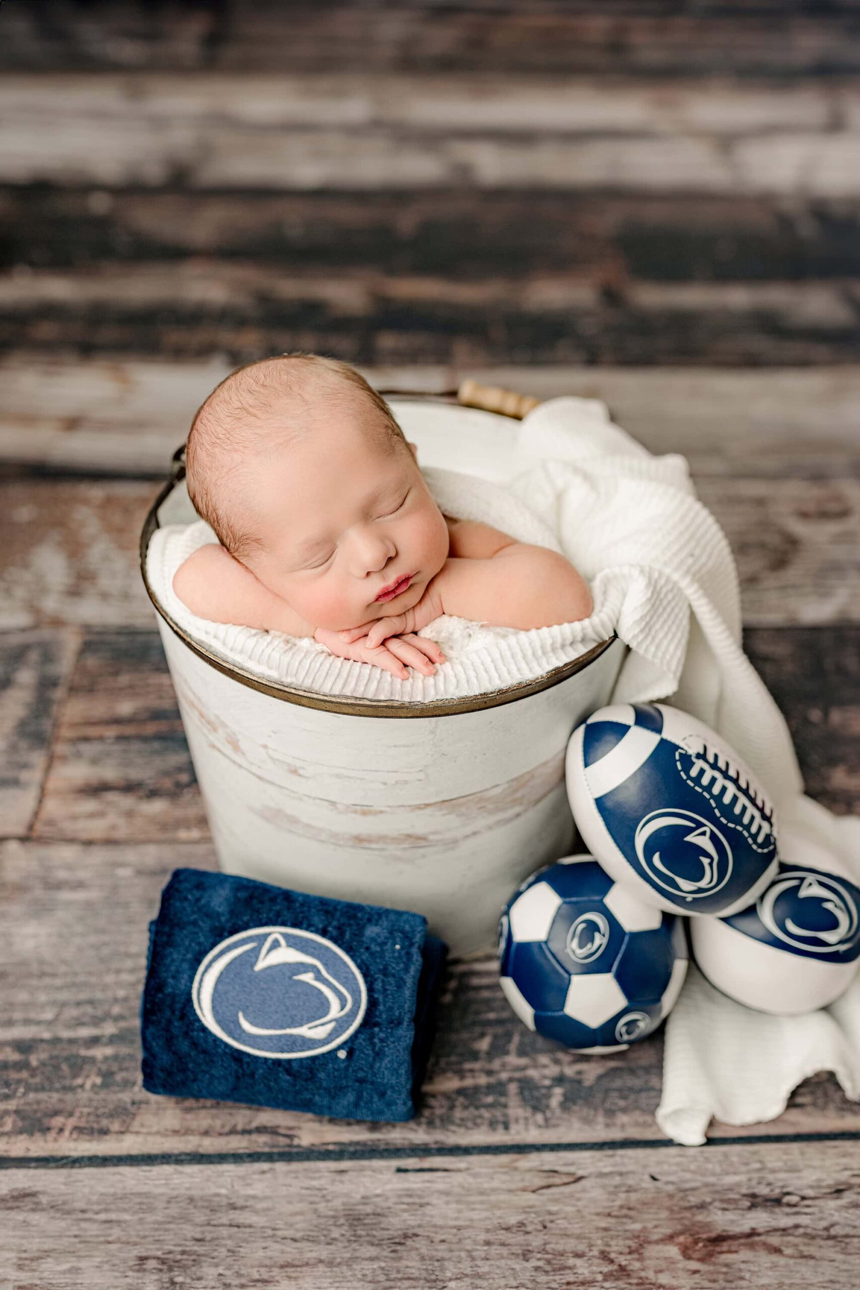 newborn baby boy posed by a specialized and trained newborn photographer in a bucket surrounded by penn state alumni gear for his penn state parents who reside in the charlotte nc area