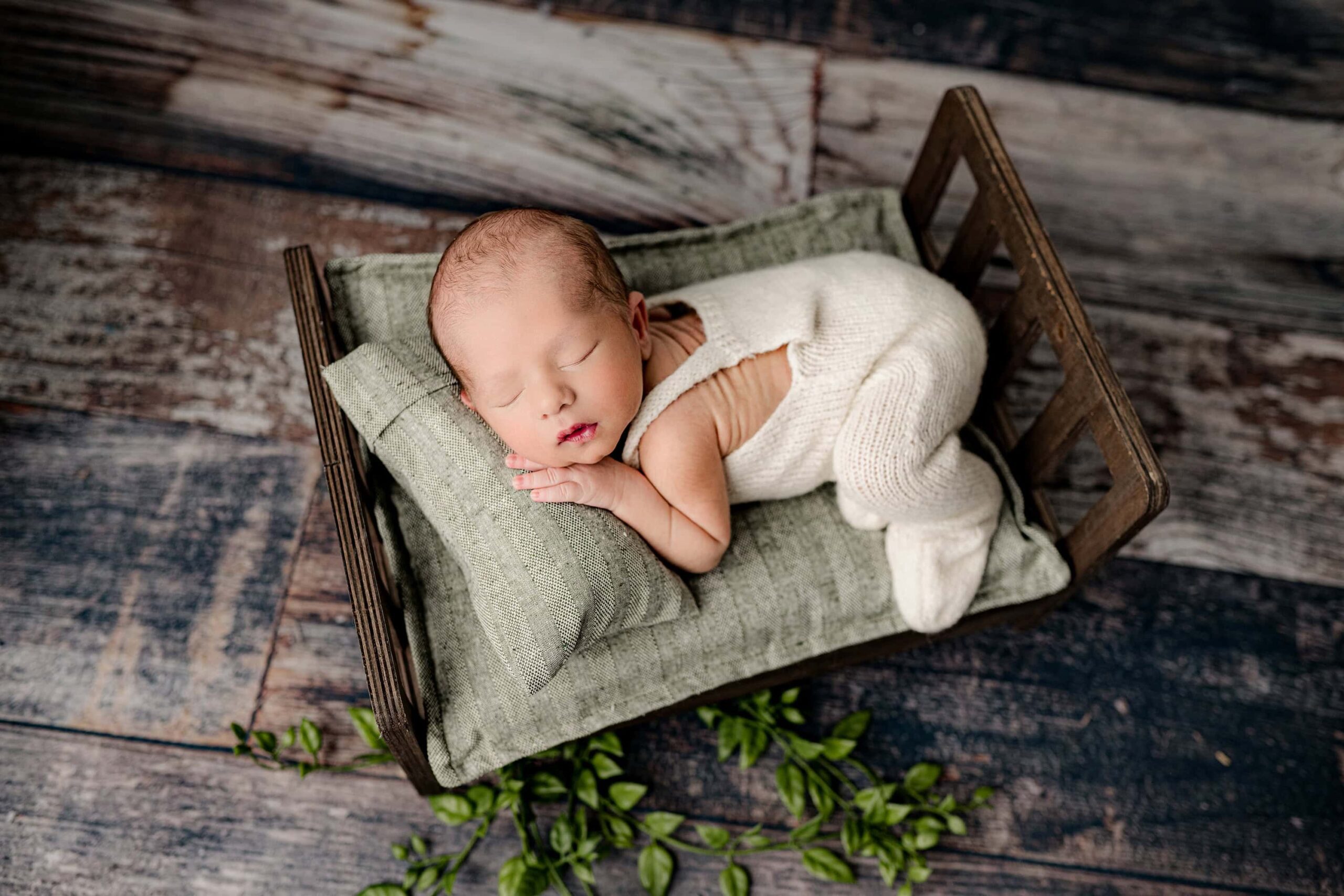 newborn boy lays on a newborn bed posed on his side during his newborn photography session dressed in white overalls and surrounded by green tones to compliment his nursery