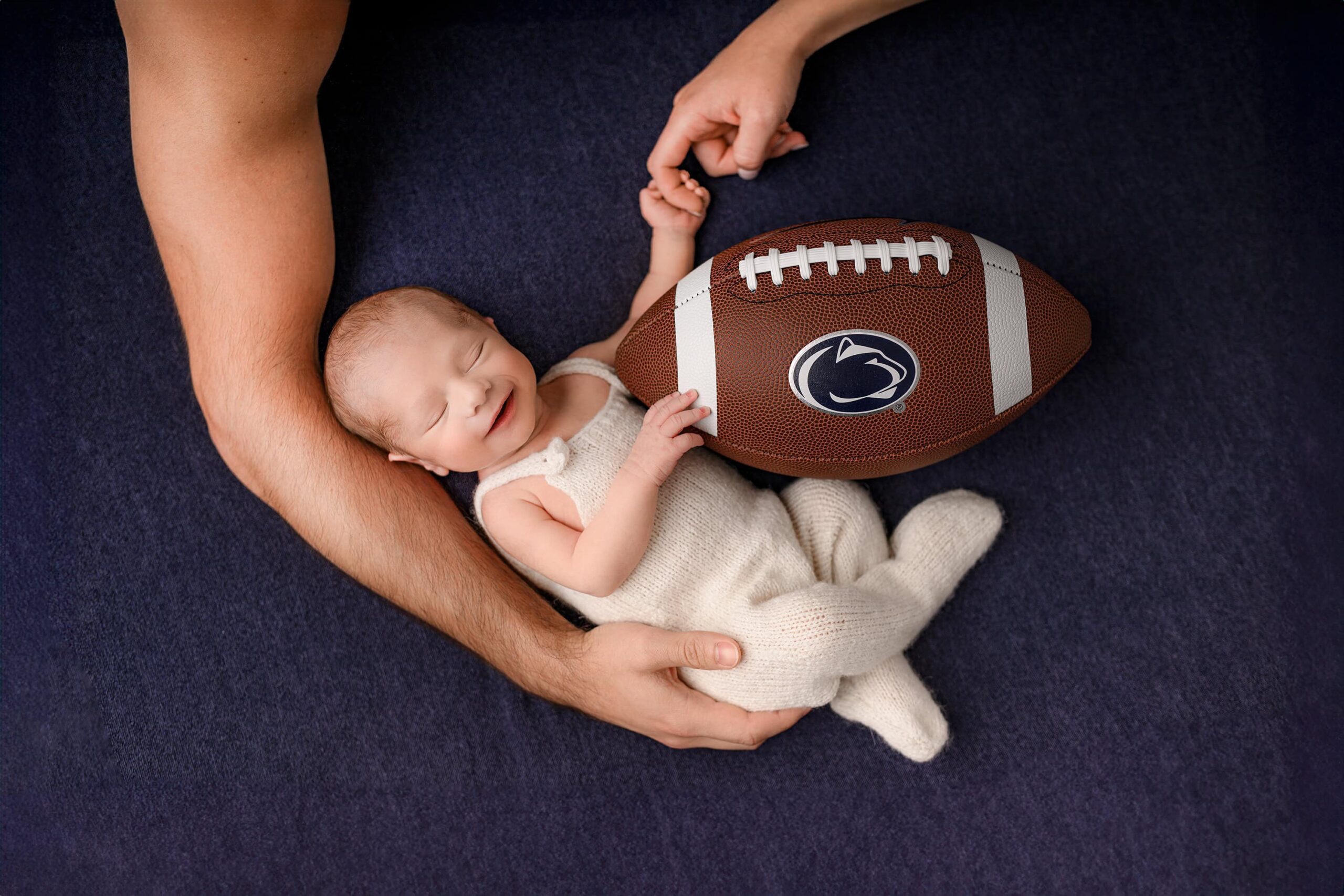 newborn baby dressed in white overalls during his newborn session with mom and dad's hands cradling him while he smiles holding a penn state football in the waxhaw, nc studio