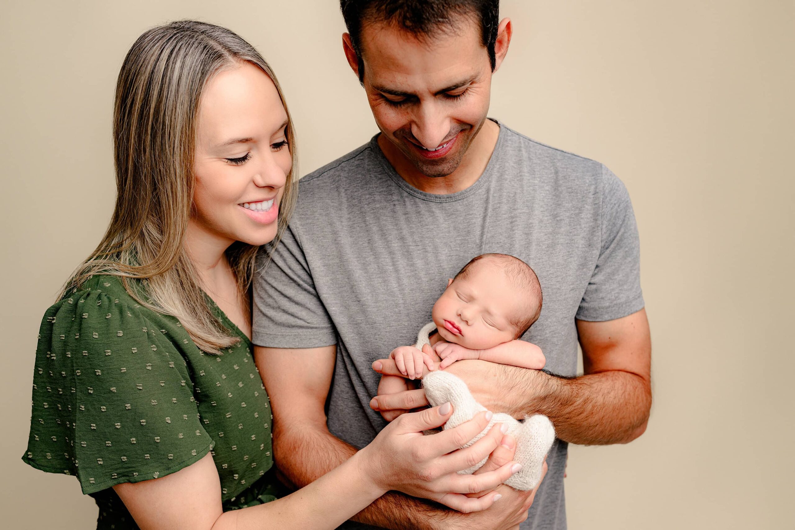 dad holds newborn son while mom and dad look down at baby smiling during their bump to baby newborn photography session in the charlotte area studio