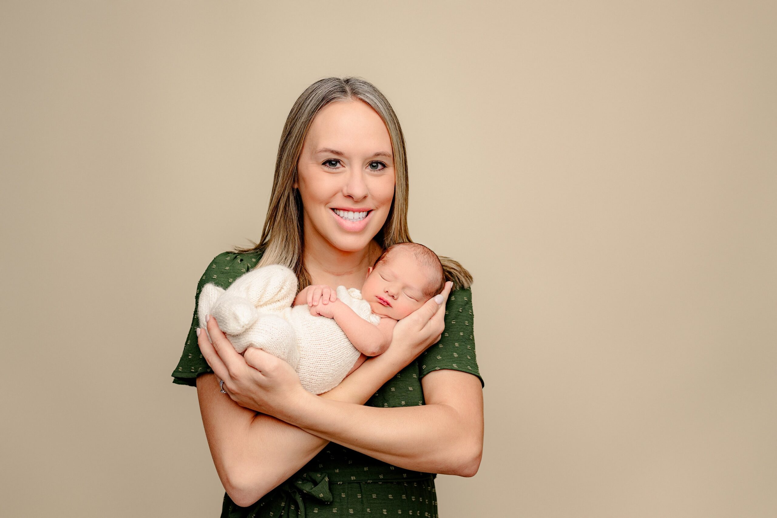 mother dressed in green gently cradles her newborn son in her arms while dressed in sweet white knit overalls in a charlotte area newborn photography studio