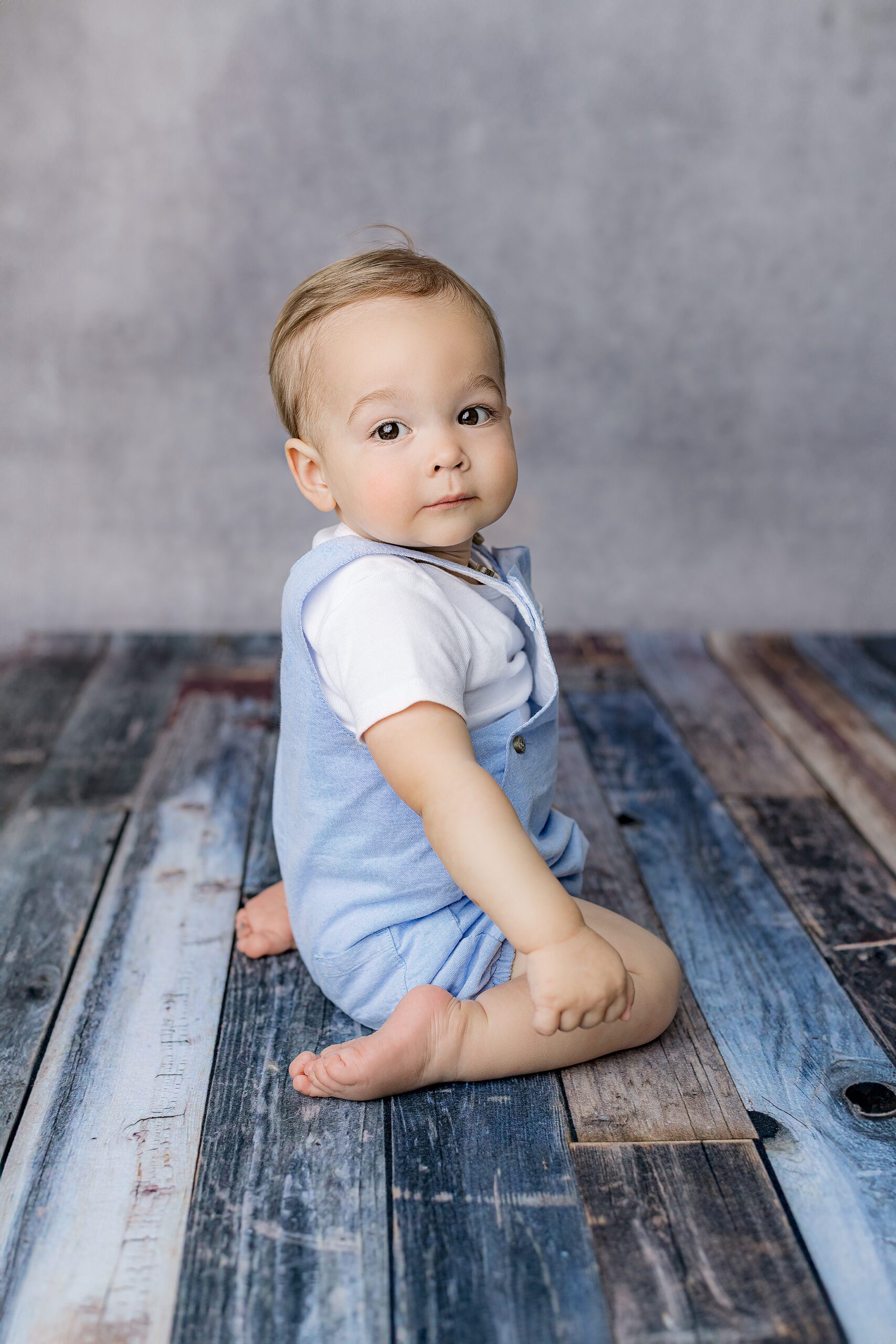 blond haired little boy sits on his knees lookin gback over his shoulder during his one year milestone session in waxhaw nc studio dressed in blue overalls and white shirt