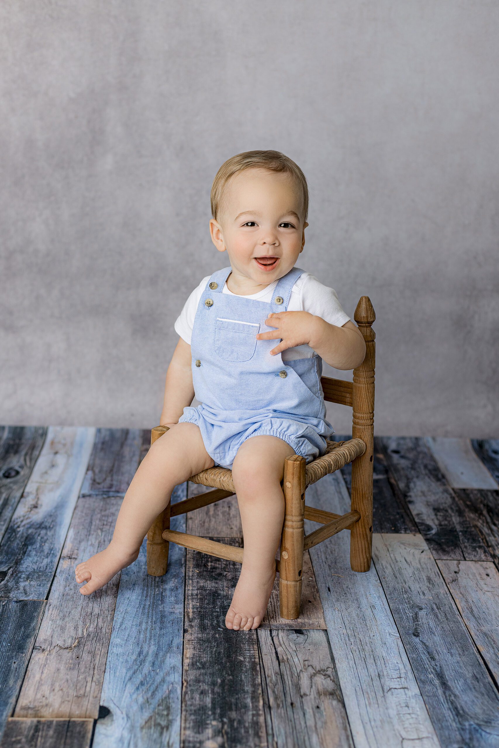 little blond hair boy laughs as he interacts with photographer during his studio milestone session to celebrate his first birthday