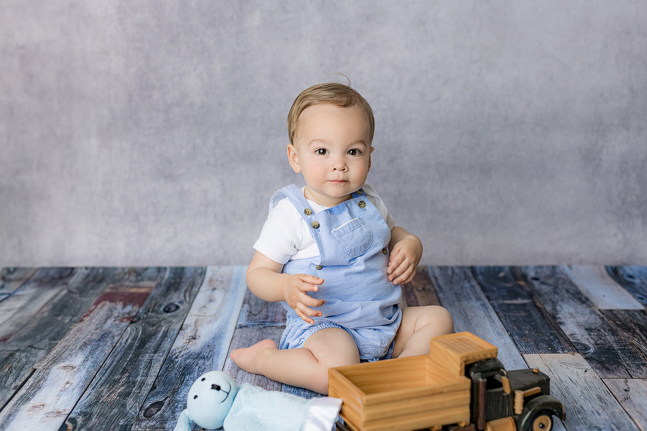 little boy sits in the blue background set up of his one year milestone session dressed in blue overalls and white shirt with a truck and lovie with him