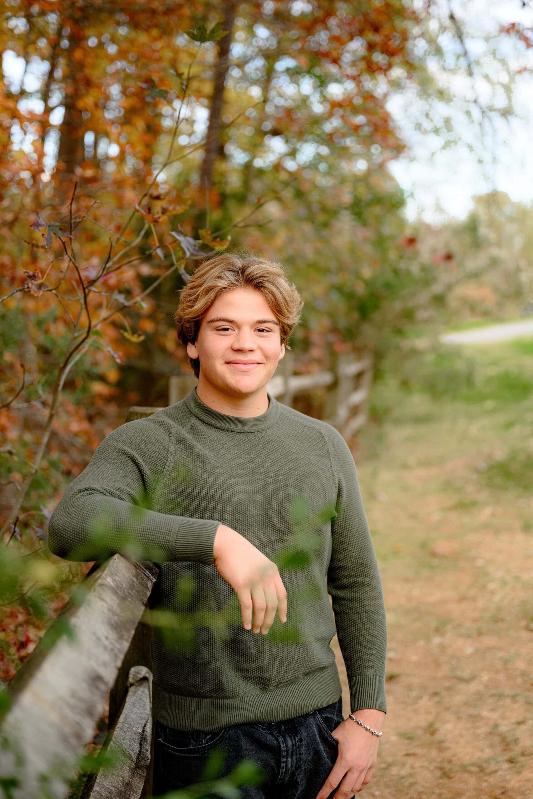 a guys stylish senior portrait sessin where the grad poses with arm up on fence class of 2025 stands wearing green sweater and dark pants