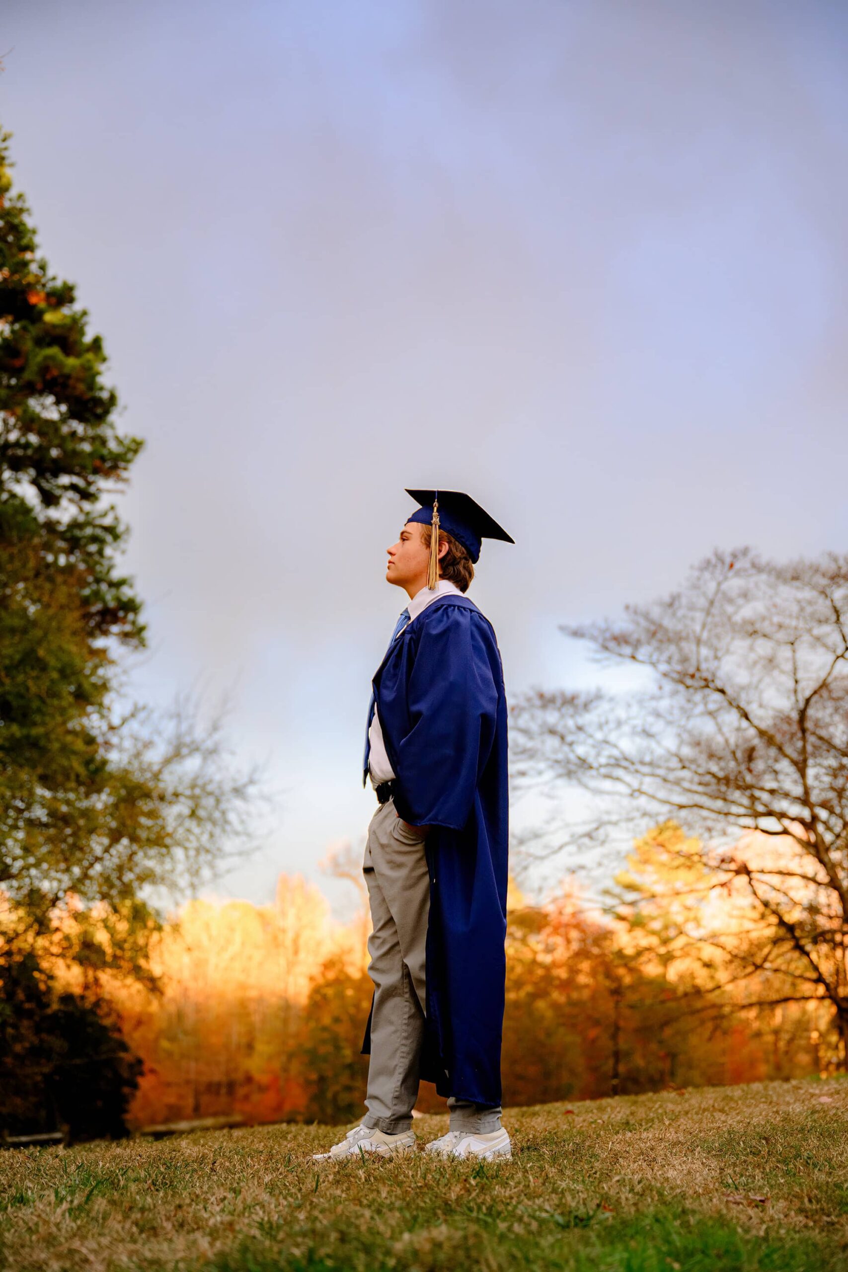 CHS and Union County grad poses for his senior portrait wearing his cap and gown and formal attire as the sun sets behind him dramatically at a waxhaw park