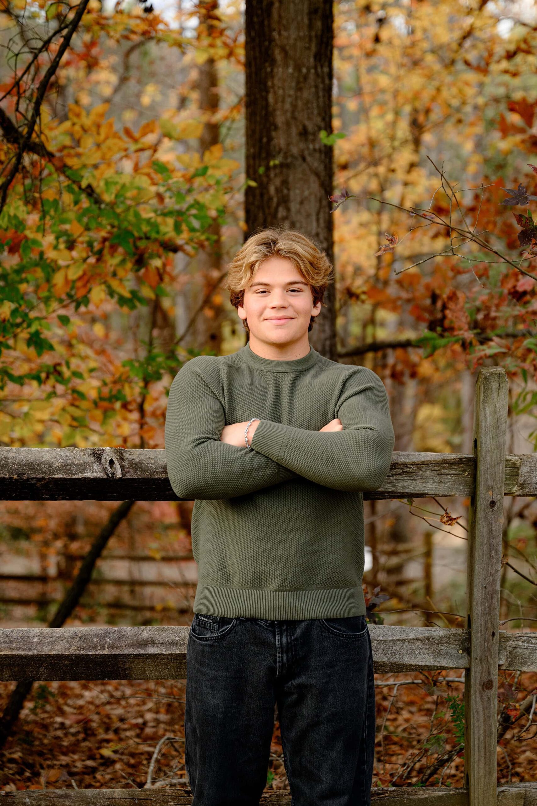 A Cuthbertson grad with his arms crossed leaning against a fence this class of 2025 grad is surrounded by fall color while wearing a green sweater and pants