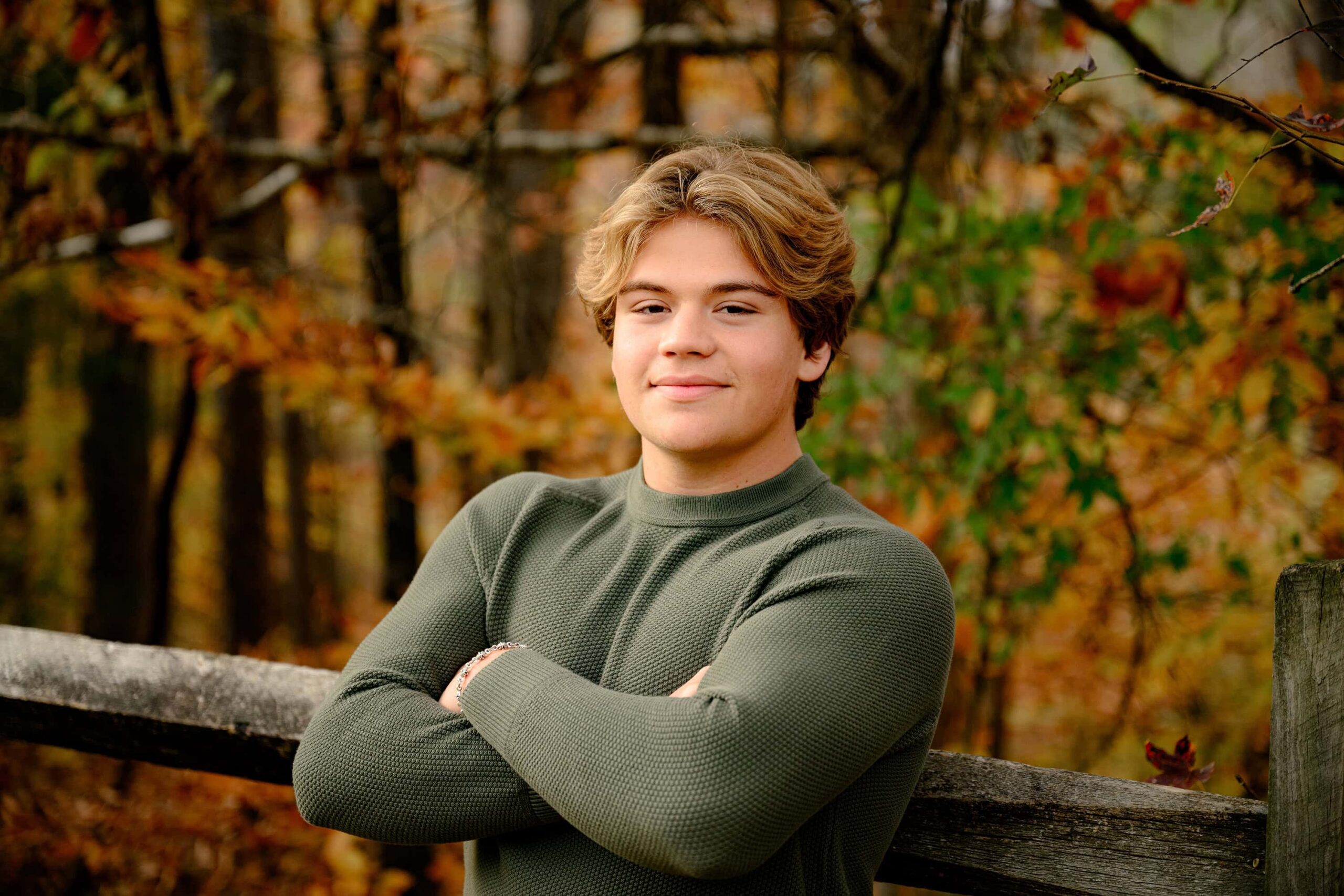Cuthbertson High School Class of 2025 senior poses with arms crossed wearing a green sweater and surrounded by fall color in waxhaw area park in his guy senior portrait session