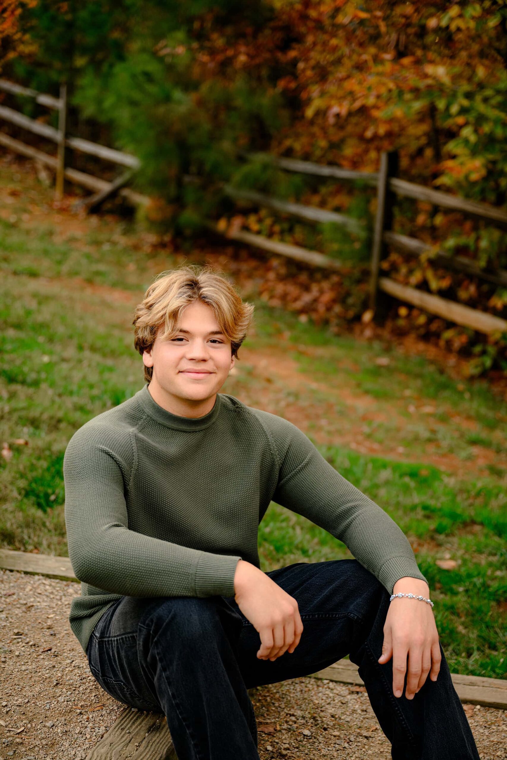 CHS grad sits wearing green sweater and dark pants surrounded by fall colors and trees showing off his style during his senior session