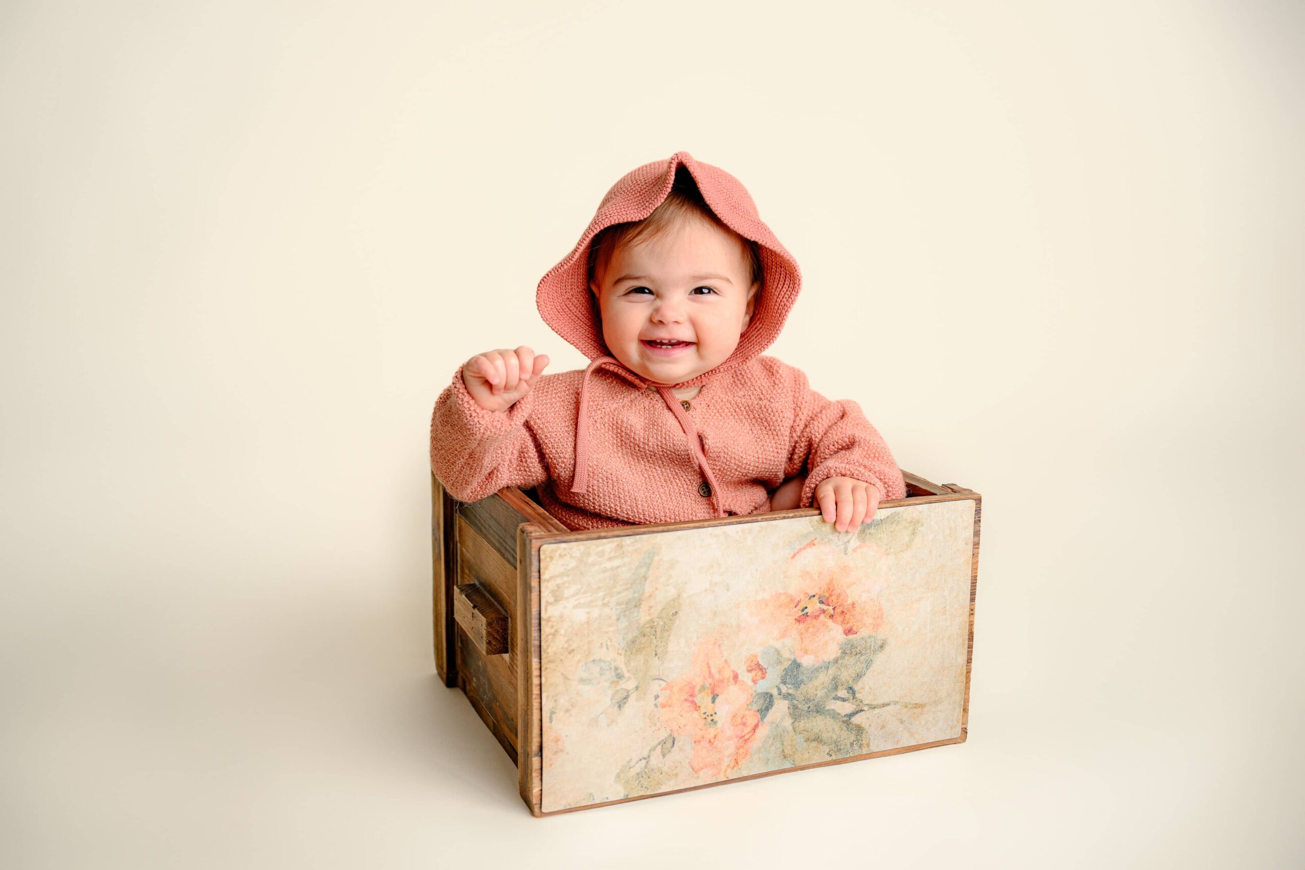 Happy one-year-old baby girl sitting in a rustic wooden crate, wearing a bonnet, smiling sweetly for her simple cake smash milestone session
