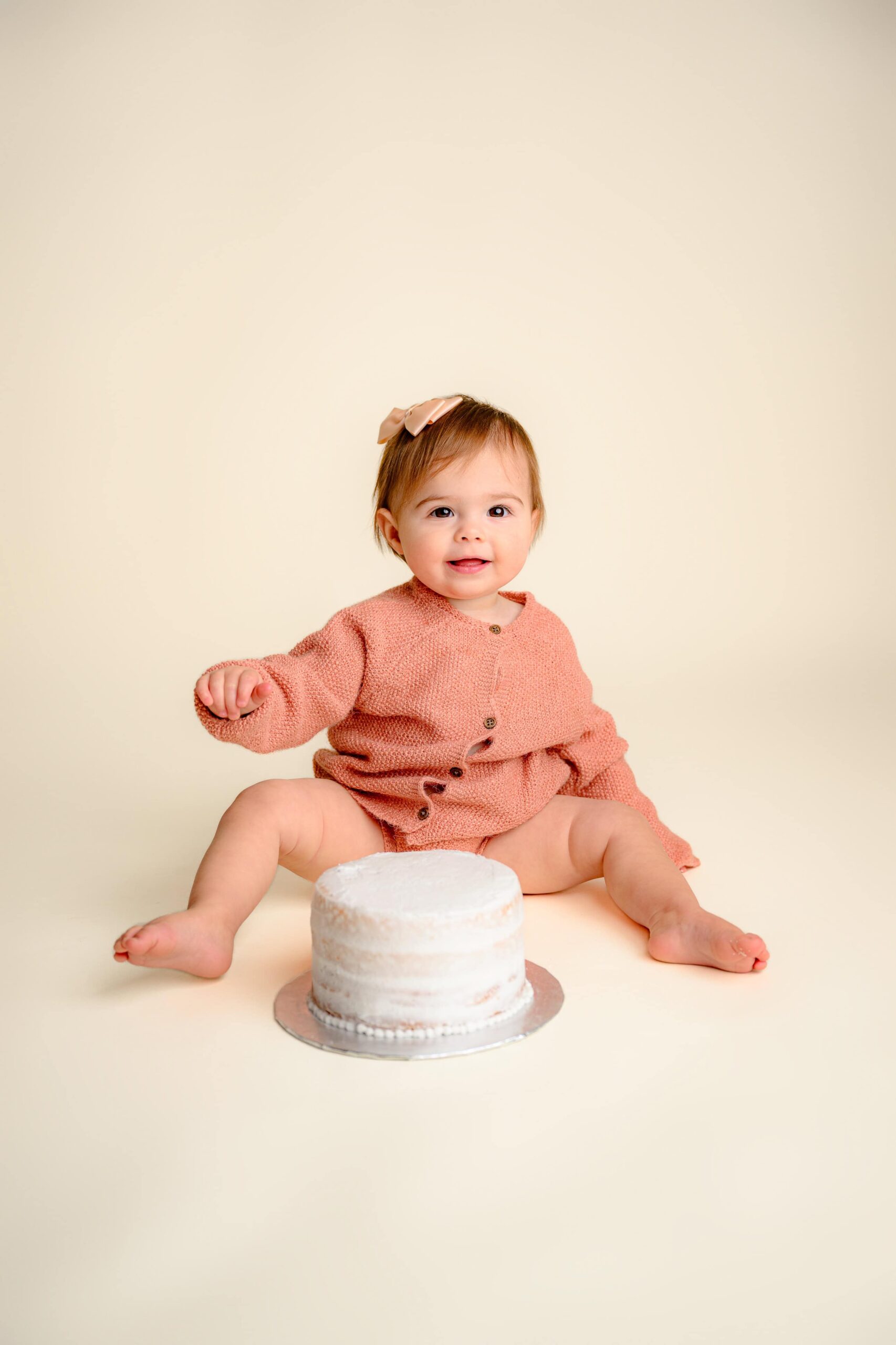 Happy one-year-old baby girl clapping sitting in front of her cake during her smash smiling sweetly for her simple cake smash milestone session