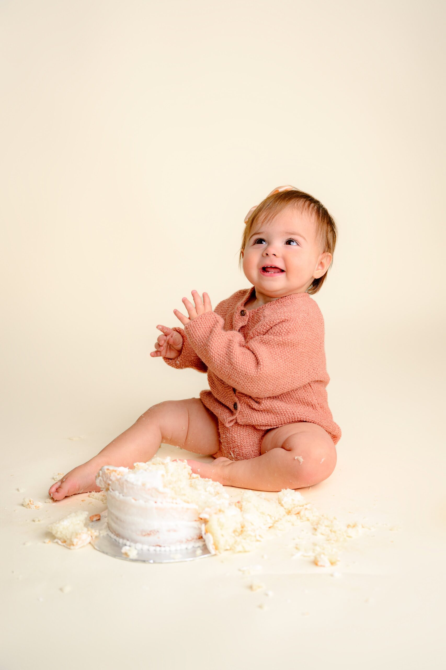 Happy one-year-old baby girl clapping and sitting beside her cake during her smash smiling sweetly for her simple cake smash milestone session