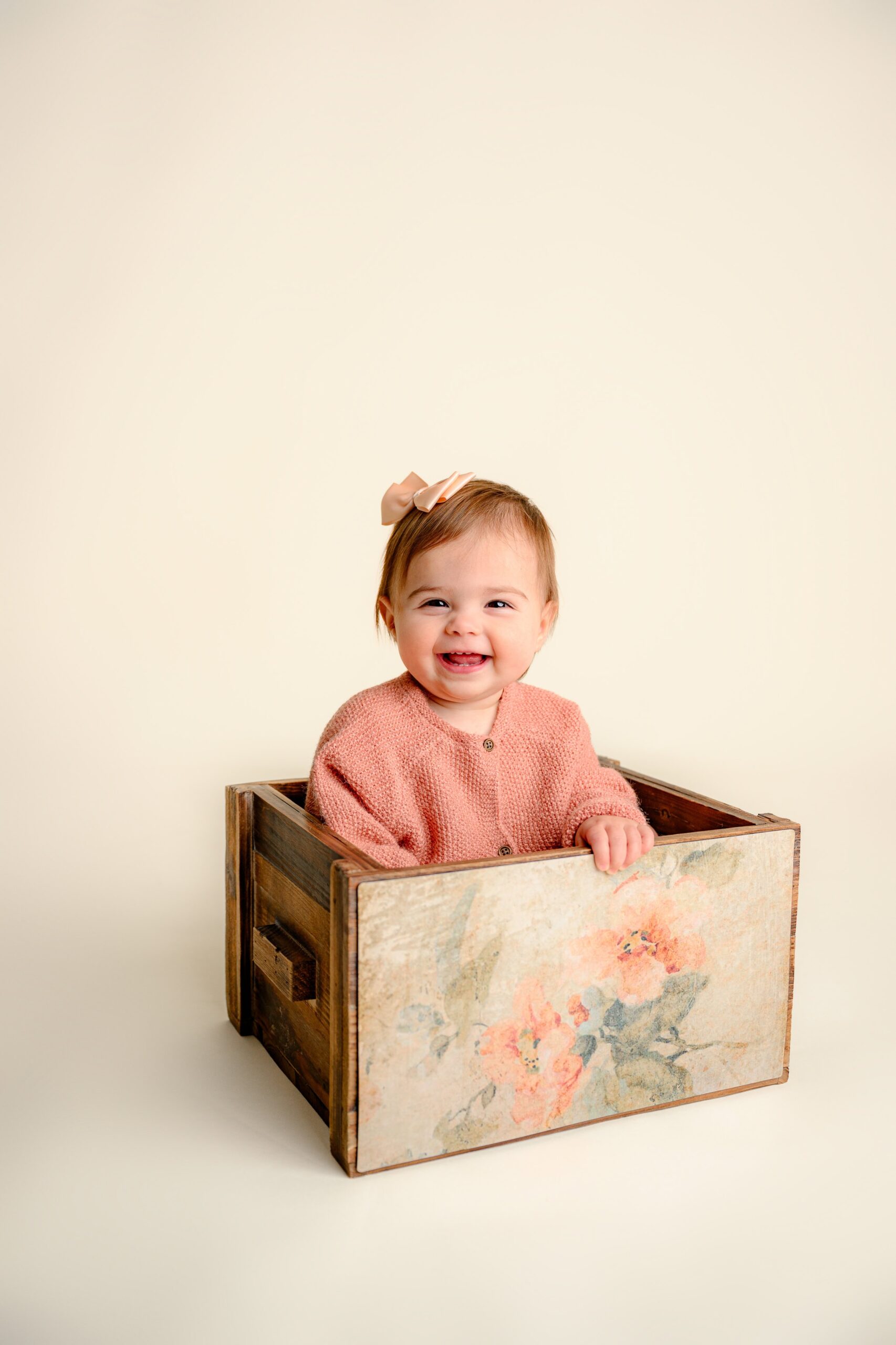 Happy one-year-old baby girl sitting in a rustic wooden crate smiling sweetly for her simple cake smash milestone session
